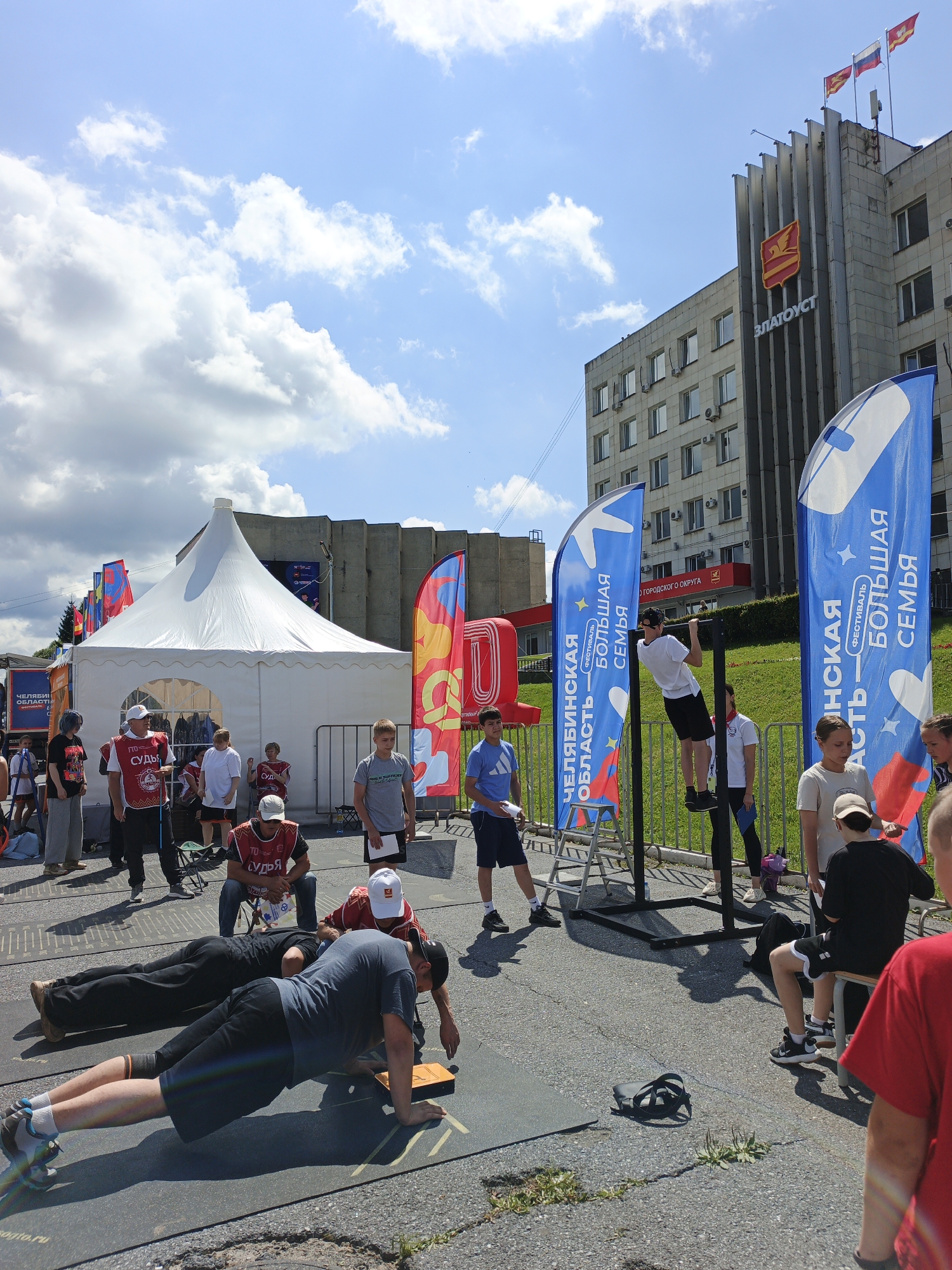 An outdoor event is underway on a sunny day in Zlatoust, Russia, with a large building in the background bearing the city's name. Several flags are displayed, including blue flags with white text that reads "CHELYABINSK REGION - BIG FAMILY FESTIVAL." A white tent is set up, likely for an event or vendor. In the foreground, several individuals are engaged in physical activities, with some performing plank exercises on mats. Others are observing or participating in the event. The ground is paved, and there are various poles and structures suggesting an organized setup for the event. The sky is partly cloudy, and shadows indicate the sun is overhead.