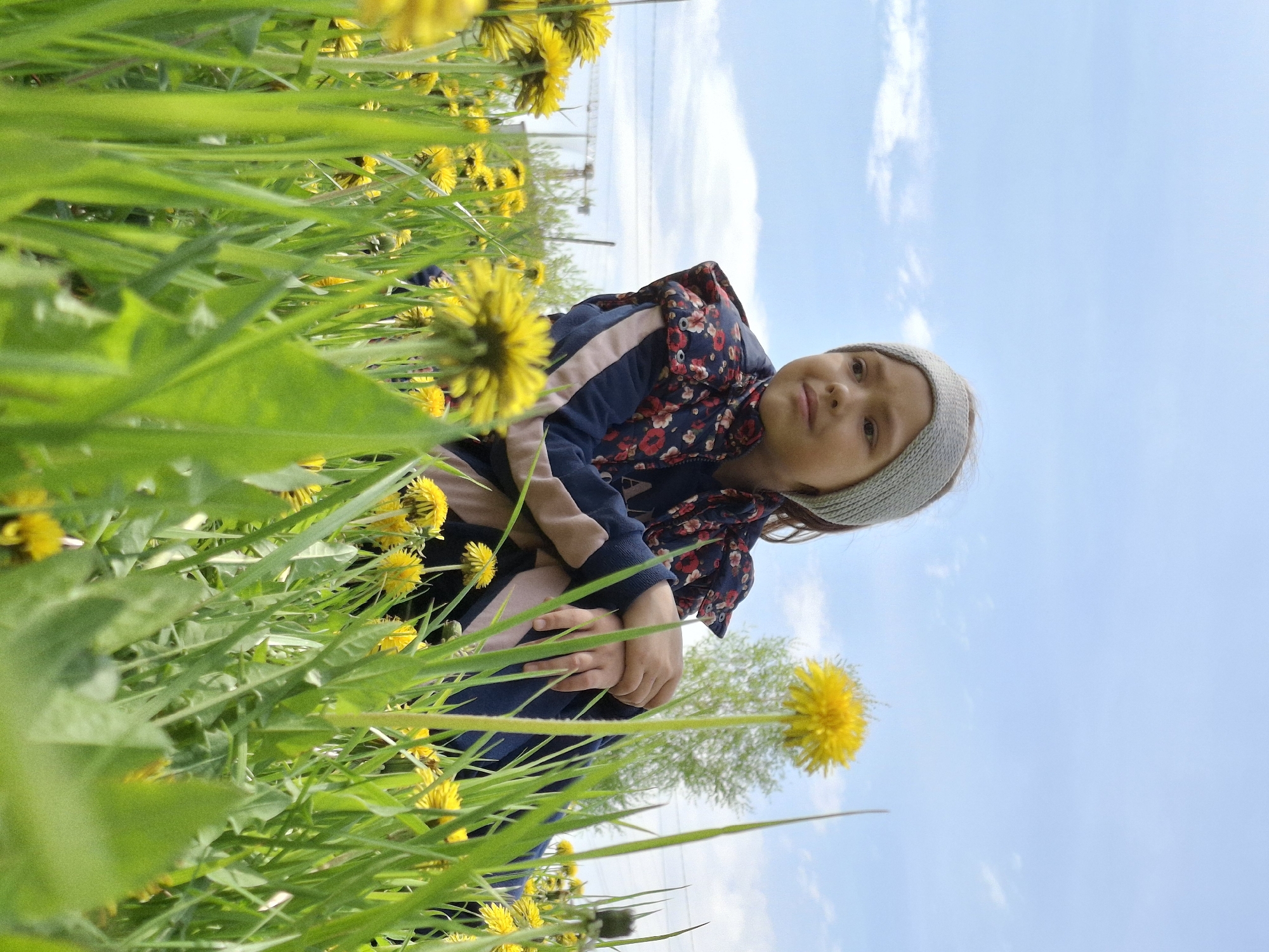 A young child wearing a dark blue jacket with a floral pattern and a light gray headband is crouching in a field of tall green grass and yellow dandelions. The camera angle is low, looking up at the child, with the sky visible in the background. The sky is blue with some white clouds. There are utility poles or structures visible in the distance. The location is possibly Chaikovsky, Russia, given the prompt.