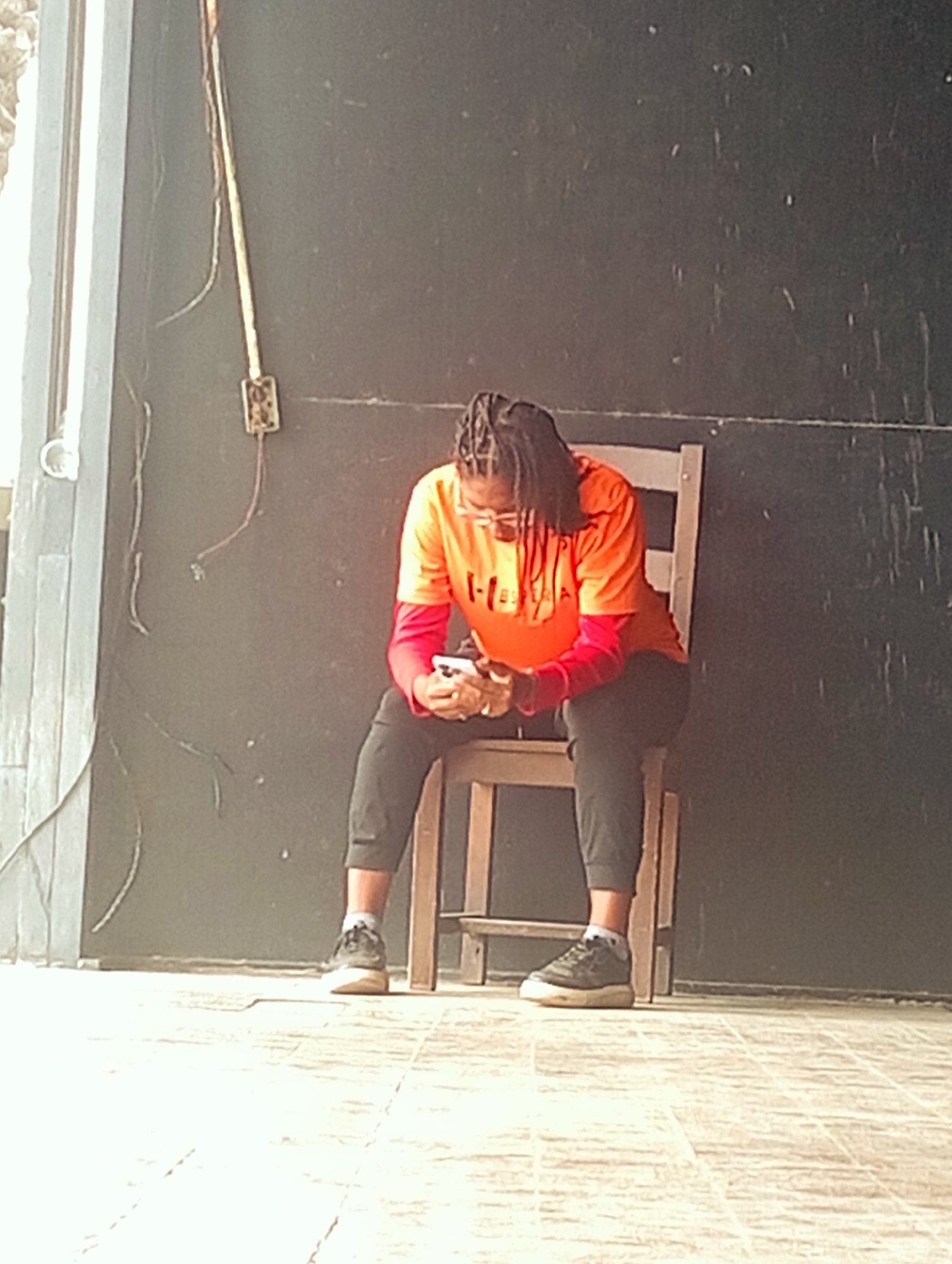 A person with dark skin and dreadlocks is sitting on a wooden chair, looking down at a cell phone in their hands. They are wearing an orange short-sleeved shirt over a red long-sleeved shirt, black pants, gray socks, and gray sneakers. The person appears to be outdoors. Behind the person is a dark, possibly painted, wall. To the left of the person is a light-colored wall with electrical wiring. The ground is covered in light-colored tiles. Based on the location provided, this scene is likely in or near Playa El Agua, Venezuela.