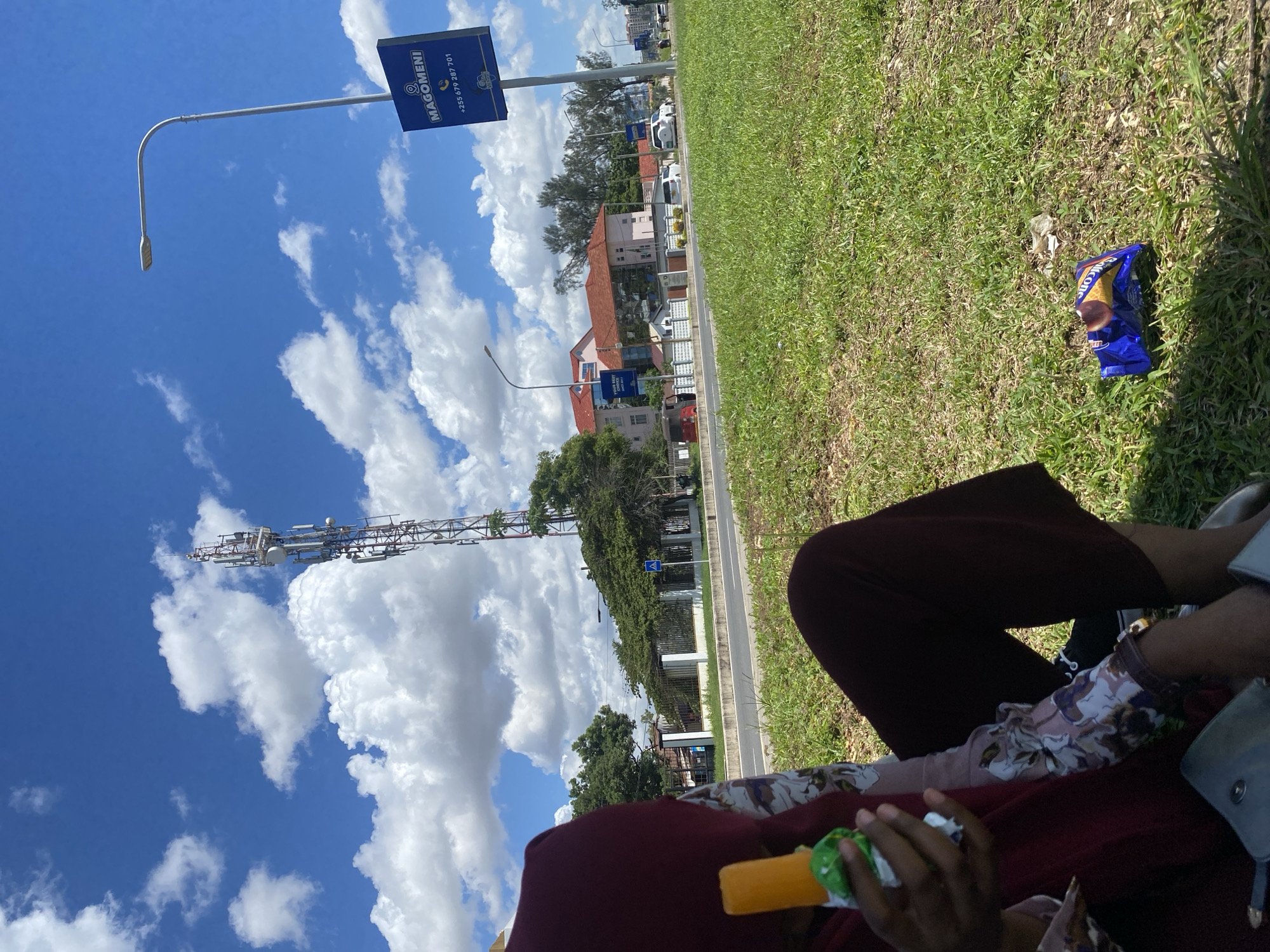 This image, which appears to be captured in portrait mode and then rotated 90 degrees counter-clockwise, depicts an outdoor scene on a clear, sunny day, likely in Dar es-Salaam, Tanzania.
In the foreground, a person is seated on vibrant green grass. Only their lower body is visible, dressed in dark maroon pants and a floral-patterned top or scarf. One hand holds a yellow popsicle or ice lolly, still partly encased in green packaging. A light blue/grey handbag rests on their lap. To the right, a crumpled blue snack bag with white text (partially legible as "RKOON") lies on the grass.
The mid-ground features a paved road with some distant vehicles. Along the road, a street light with a long, curved arm extends from the left. A tall, metal cell tower, equipped with multiple antennas, prominently stands against the sky, visible above the road and trees. Further back, several buildings with reddish-brown roofs are nestled among green foliage.
The background is dominated by a bright blue sky dotted with fluffy white cumulus clouds. A blue rectangular sign on a pole, situated by the road, displays white text that reads "MAGOMENI" and a phone number: "+255 679 287 701". The overall impression is a casual, relaxed moment spent outdoors in an urban or suburban environment during daytime.