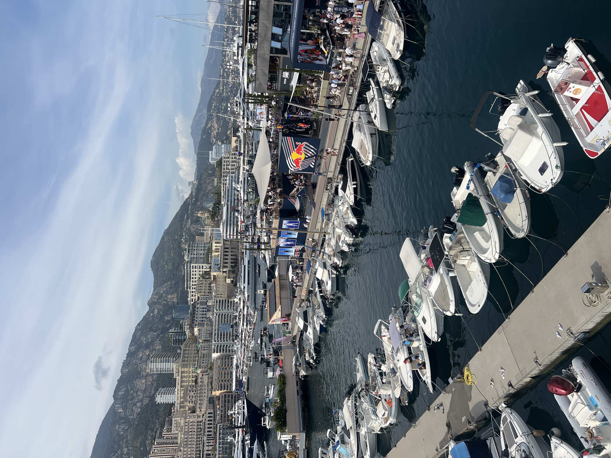 This high-angle shot captures Port Hercule in Monaco, showcasing a bustling marina nestled against a backdrop of densely built hills. In the foreground, several white motorboats are moored alongside a concrete jetty, with mooring lines and fenders visible. Some boats have small canopies or covers, and scattered items like yellow hoses and white flip-flops are on the jetty.
The midground features a wider section of the port with numerous larger yachts and motorboats, including several superyachts, populating the deep blue water. Along the main quayside, temporary hospitality structures are erected. A prominent two-story structure displays a large Red Bull logo with racing-themed graphics, and what appears to be a replica or display of a race car is integrated into its design. Crowds of people are visible on the quayside, congregating around these structures and walking along the waterfront. Another structure with "VISA PR" branding is also present. A large white tent provides shade over a gathering area.
The background reveals the city of Monaco ascending the steep, rocky, and heavily vegetated hillsides. A mix of architectural styles is apparent, from classical ornate buildings to modern high-rise towers. Further in the distance, additional mountains fade into the hazy blue sky, which has scattered wispy clouds. The overall scene suggests a vibrant event or peak tourist activity under bright daylight.
