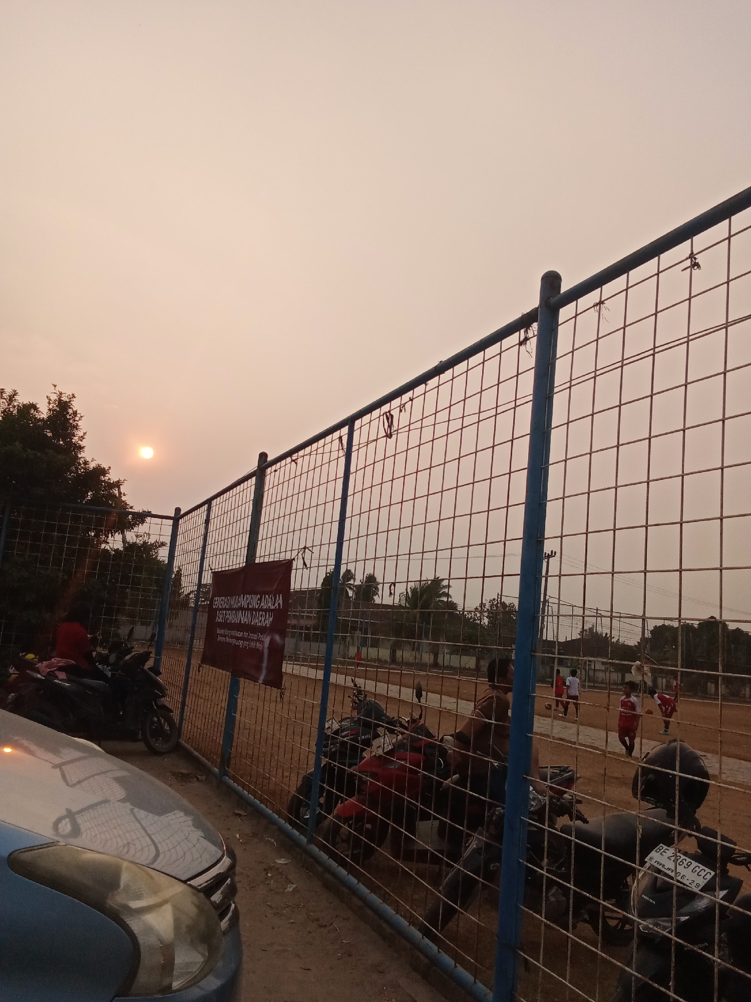 The image depicts an outdoor scene at what appears to be a sports field, likely in Toto Katon, Indonesia, during a hazy sunset. A tall, blue-railed fence with a wire mesh divides the foreground from the background. On the left, a light blue car is parked, its hood reflecting the fence and sky. Behind the car, several motorcycles are parked, some with riders.
The field itself is a dirt surface, and several people are visible, some wearing red and white athletic attire, suggesting a game or practice is in progress. There are trees and buildings in the distance, partially obscured by the haze and the fence. A maroon banner with Indonesian text is attached to the fence. The sun is low in the sky, casting a warm, diffused light over the entire scene. The overall impression is one of an active community space at the end of the day.