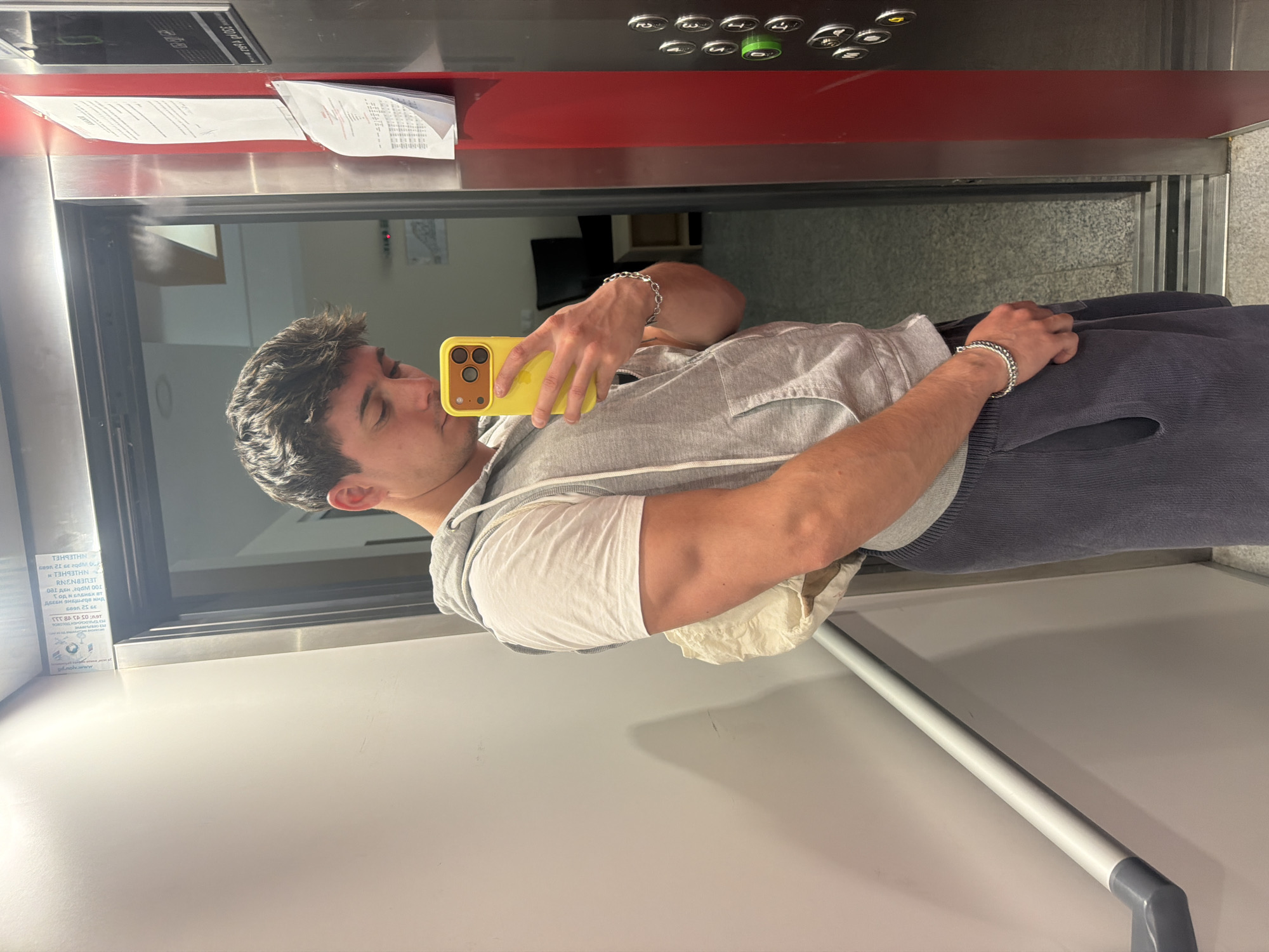 A young man is captured in a mirror selfie inside an elevator, likely located in Sofia, Bulgaria. He appears to be in his late teens or twenties, with short, dark, textured hair. He is dressed casually in a light grey hooded sweatshirt layered over a white t-shirt and dark blue trousers, possibly corduroy. A silver-colored chain bracelet adorns his left wrist. He holds a bright yellow smartphone, featuring a prominent triple camera setup, in front of his face to take the picture. His eyes are closed, suggesting a posed shot or a momentary expression.
The elevator's interior is reflected in the mirror. Above the man's reflection, a red control panel with silver buttons is visible, including numbers "3" and "2," and a glowing green button. Papers are tucked into a narrow shelf directly above the control panel. Visible text on these papers includes "3952 1609" and Cyrillic script such as "ГОД И БЛЕНЕХ ВТ" and "081 ден 290 001," which aligns with the provided location context of Bulgaria.
On the reflected left wall of the elevator, a white sign or sticker displays reversed text. OCR deciphers parts of this as "THN ODRPATSKG HASY," "LEVEBNINY," and "NHIEUHEL," possibly indicating local information or advertisements within the building. A white handrail extends across the bottom of the reflected scene. The reflection in the elevator doors shows an interior hallway or room.