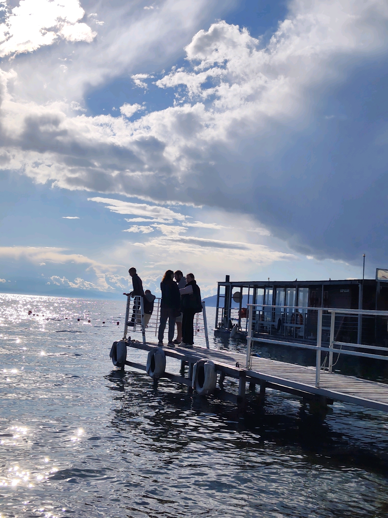 A group of five people are standing on a wooden pier extending into a body of water. The pier has white railings and is supported by pilings. The water is choppy and reflects the sunlight, creating a sparkling effect. In the background, there is a floating structure with large windows, possibly a cafe or restaurant. The sky is filled with dramatic clouds, some bright and white with sunlight breaking through, others dark and ominous, suggesting an approaching storm. The overall impression is a moment of contemplation or conversation on a pier, with a striking sky and water backdrop. The location is the Listvyanka settlement, on the shore of Lake Baikal in Russia.