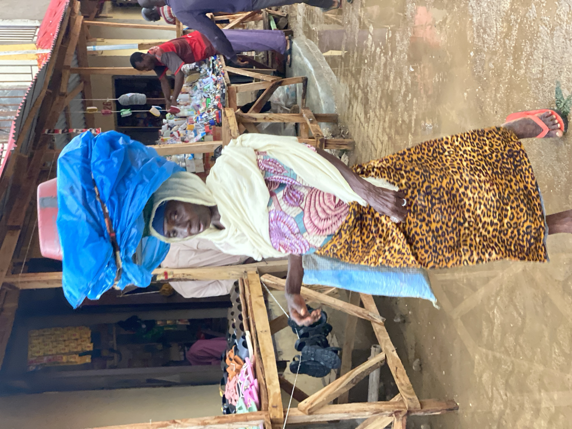 In Kafanchan B, Nigeria, a woman wearing a leopard print skirt, a patterned top and a white head covering, is walking through what appears to be a flooded area. She has a blue bag and a pink container balanced on her head. In her right hand, she holds a string with black sandals attached. Nearby, there is a structure with wooden supports where additional sandals are on display for sale. Further in the background, a man wearing a red shirt can be seen near some plastic containers, and water is visible on the ground.