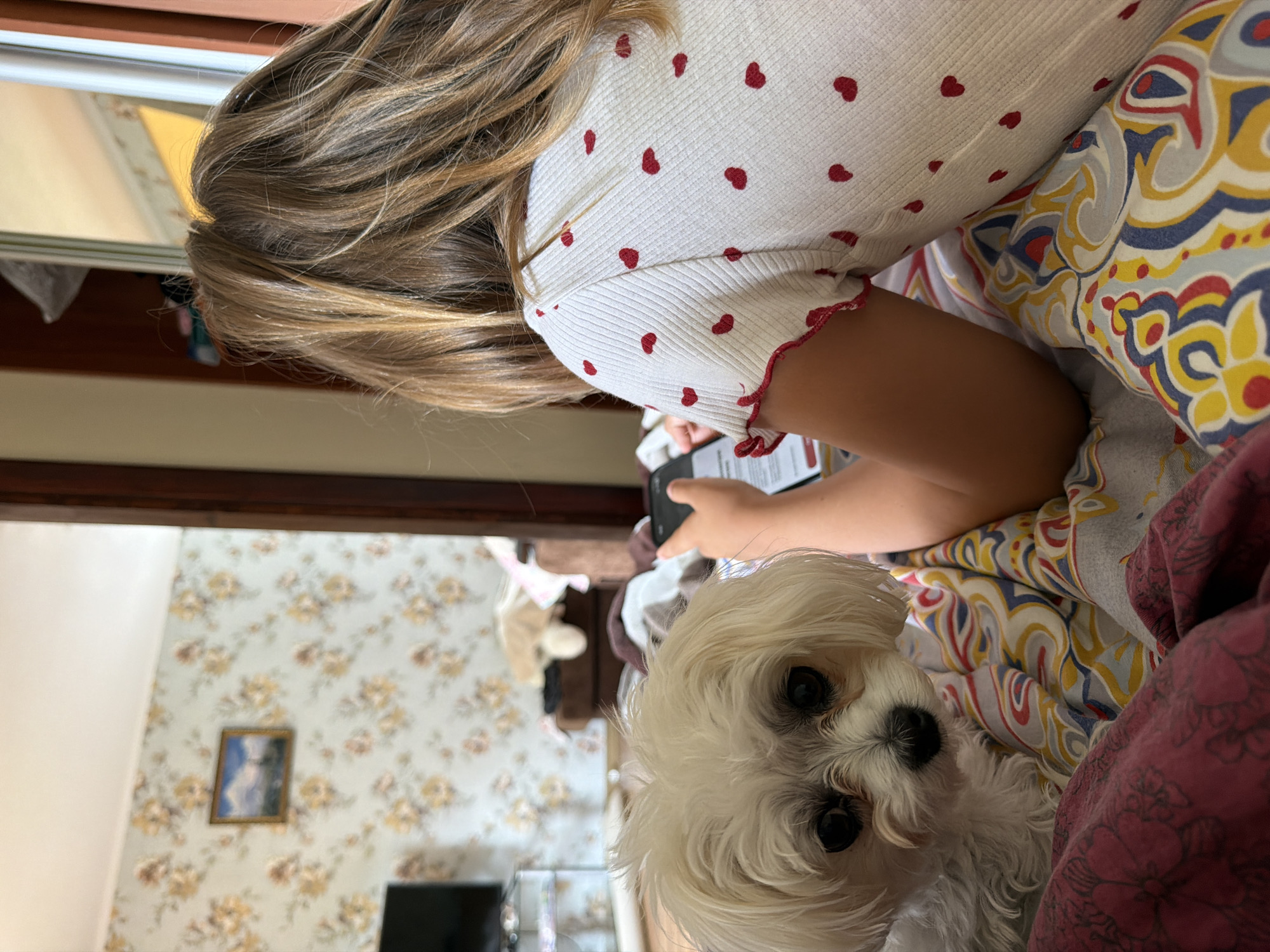 In a room in Partenit, Ukraine, a young girl with blonde hair lies on a bed, looking at her phone. Beside her is a small white dog with dark eyes. The girl is wearing a white shirt with red hearts. The bed is covered with a patterned blanket. In the background, a wall with floral wallpaper is visible, along with a framed picture. There is also a glimpse of a mirror reflecting part of the room.