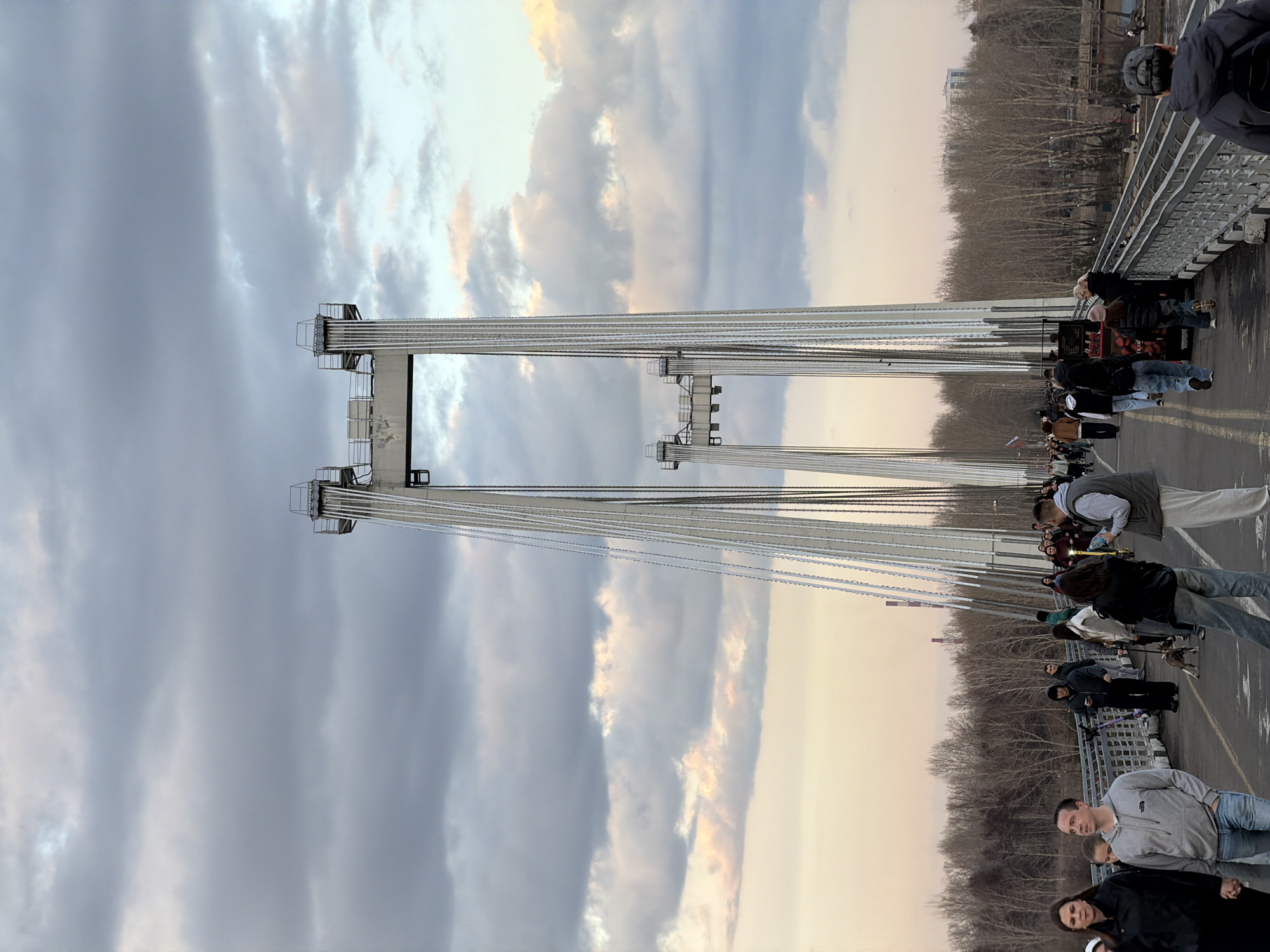 This image captures a view of the pedestrian bridge in Krasnoyarsk, Russia, with multiple people strolling across it. The bridge's towers and suspension cables dominate the scene, ascending into a sky filled with clouds, suggesting either sunset or sunrise conditions. People of various ages and attire are walking across the bridge, some appearing to be tourists or locals enjoying the view. The bridge's structure is prominent, demonstrating its architectural design and engineering. On both sides of the bridge, trees are visible, possibly bordering the river or park area.