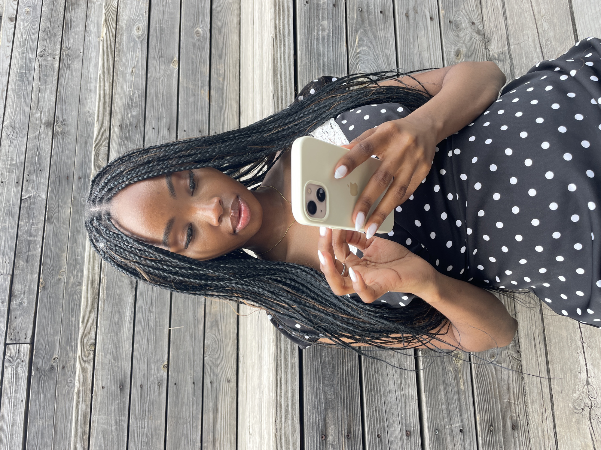 In Kazan, Russia, a young woman with braided hair is seated on wooden bleachers. She is wearing a black dress with white polka dots. She is holding and looking at a smartphone in her hands. The background consists of weathered wooden planks, likely part of an outdoor seating area or structure.