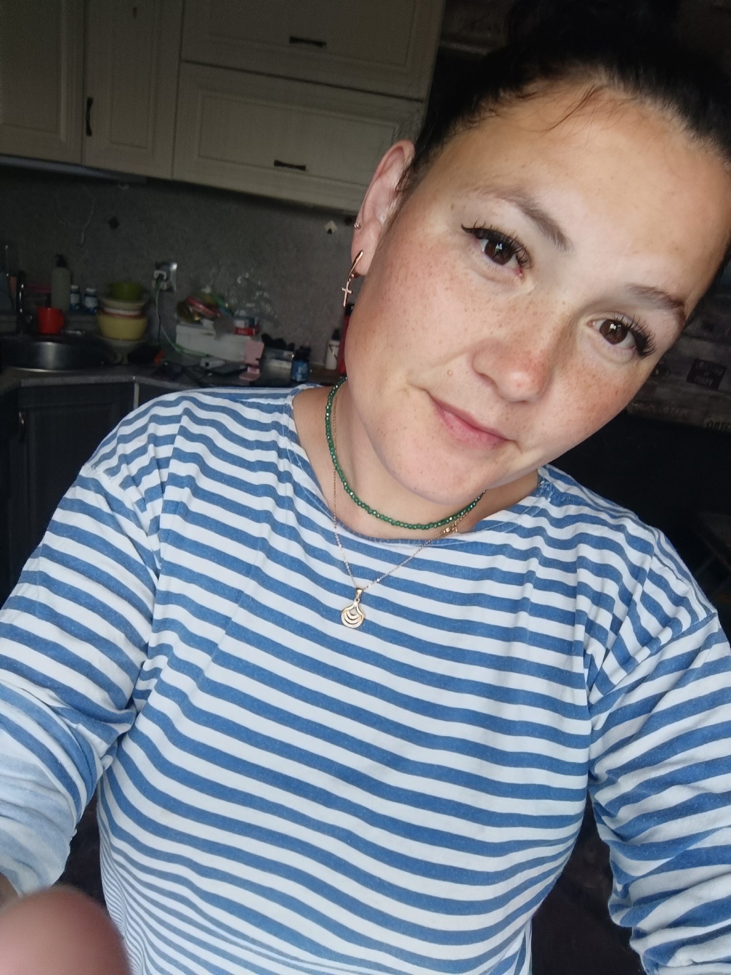 A woman with dark hair pulled up and freckles across her face is taking a selfie in a kitchen. She is wearing a blue and white striped long-sleeved shirt. She has on a green beaded necklace and a gold pendant. Behind her, kitchen cabinets and countertops are visible. The location is not specified to be in Karsha, Russia.