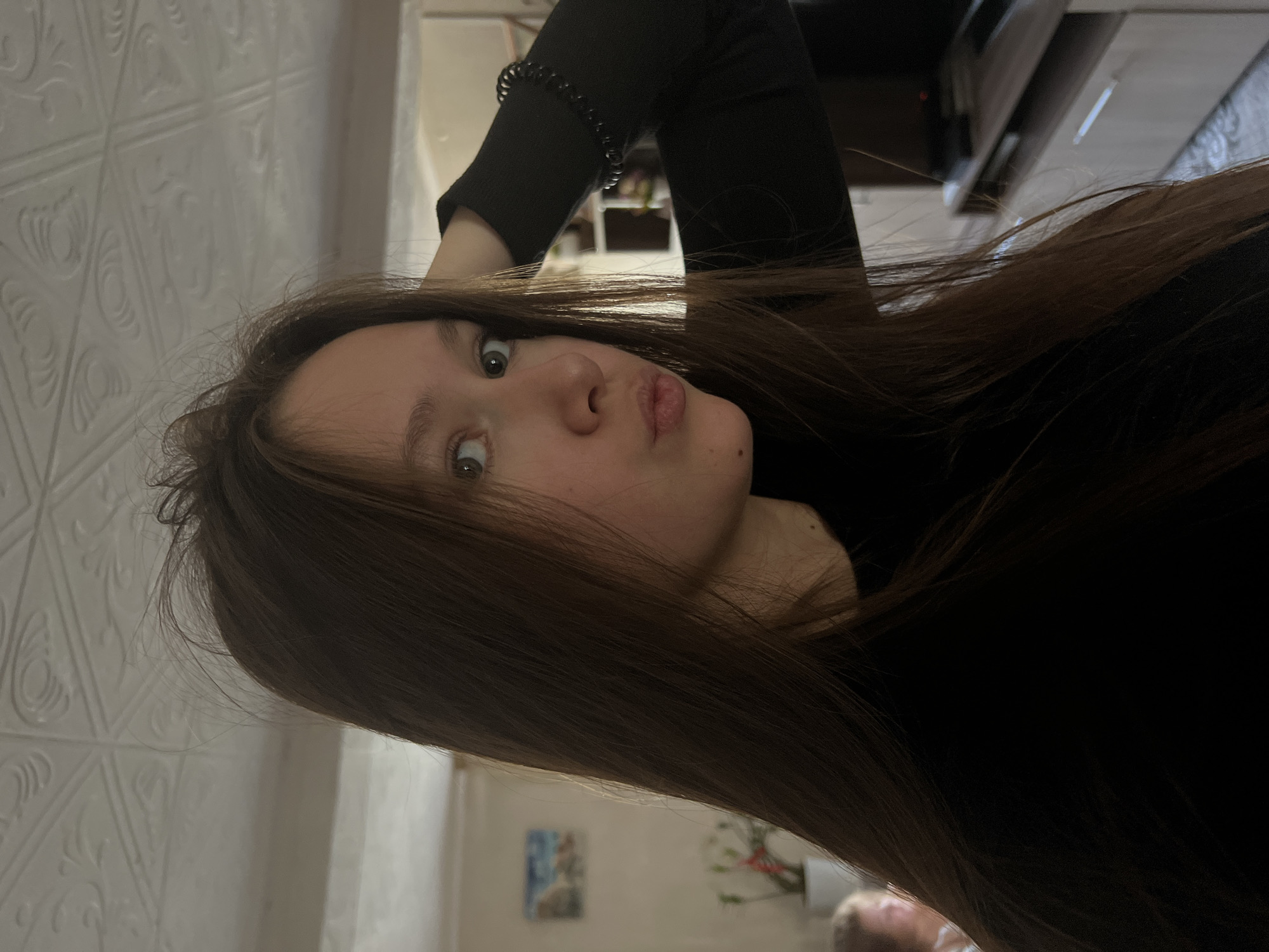 A young woman is pictured from a close, canted angle indoors. She has long brown hair and is wearing a black top. Her expression is playful, with slightly widened eyes and pursed lips. A wall with a textured, patterned surface is visible in the background. A hair tie is visible on her wrist. In the background, a picture hangs on the wall and a plant is partially visible. The location appears to be a private residence. The city, Chaikovsky, Russia, cannot be specifically verified based on visual information alone.