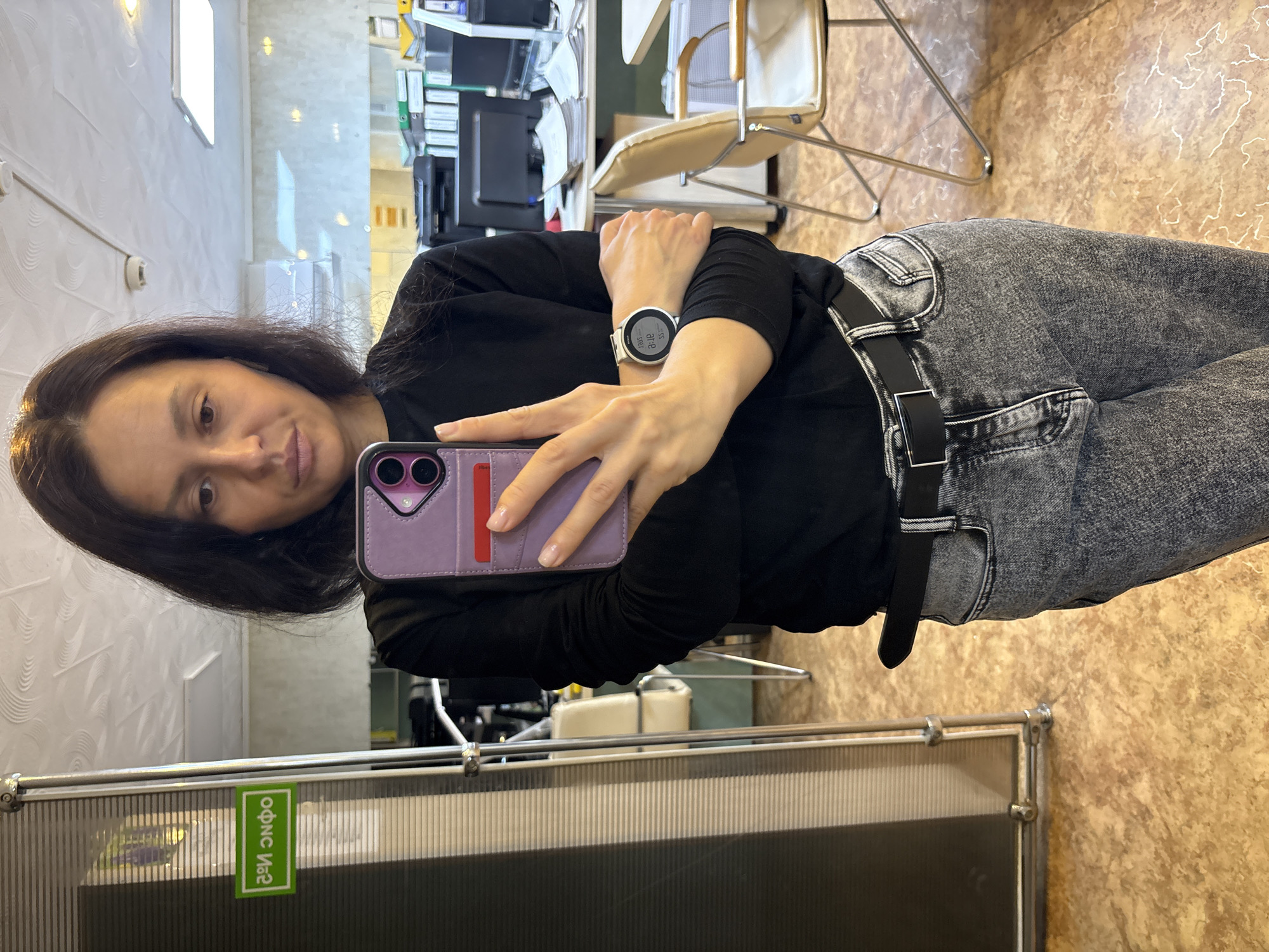 A woman is taking a selfie in what appears to be an office setting, possibly in Chaikovsky, Russia, indicated by the text "офис Ив2" (office Iv2) on a sticker. The woman is holding a phone with a purple case. She is wearing a black long-sleeved shirt and grey jeans with a black belt. She also has a wristwatch on her left wrist. The background includes office furniture, such as a desk, chair, and computer equipment. The floor has a patterned surface.