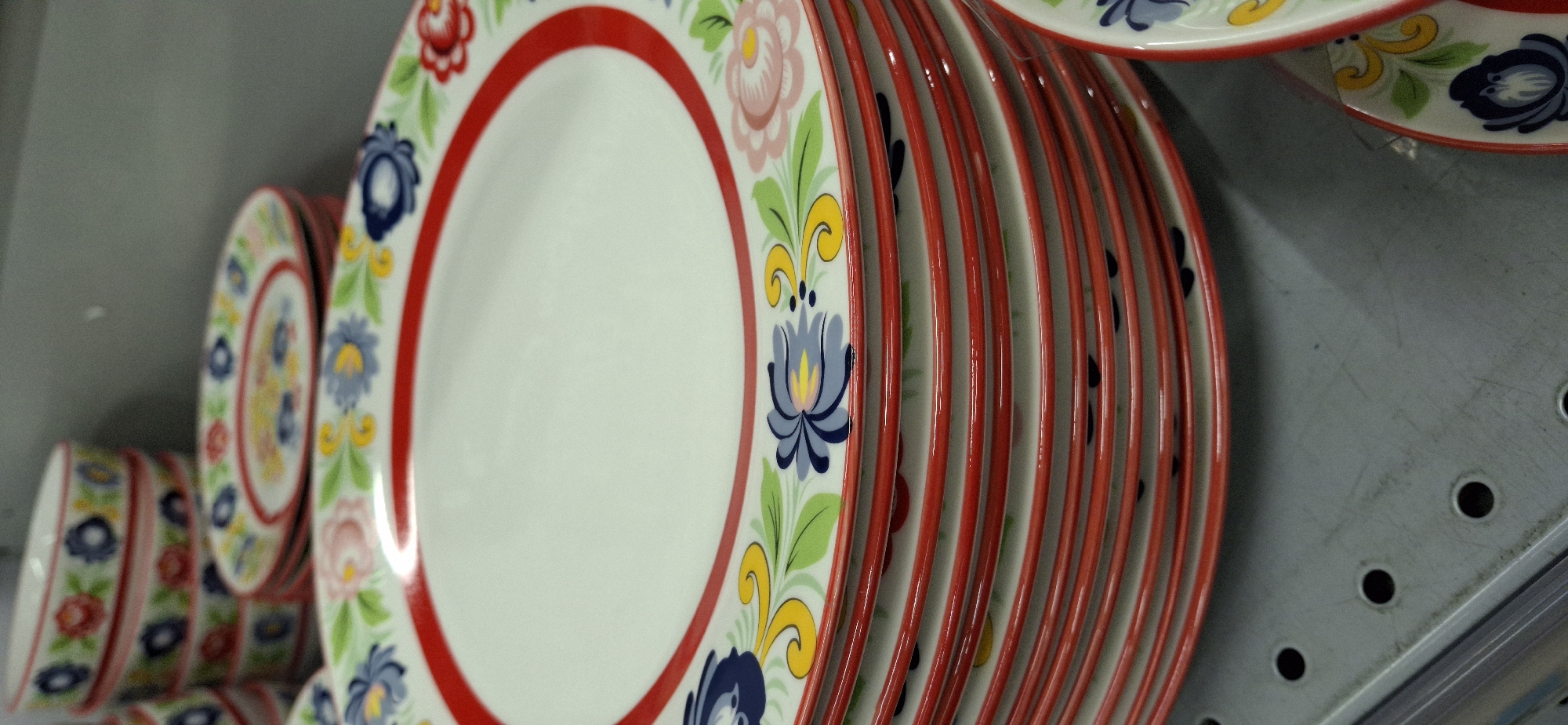 A stack of white ceramic plates with red rims and colorful floral patterns is displayed on a gray metal shelf. The plates are decorated with blue, pink, yellow, and green flowers and leaves. The shelf has circular perforations. In the background, a stack of white ceramic bowls with similar floral patterns is visible. To the right, a portion of another plate with a red rim and floral design is visible. The scene appears to be a retail display, likely in a store. The location is Telmana, Russia.