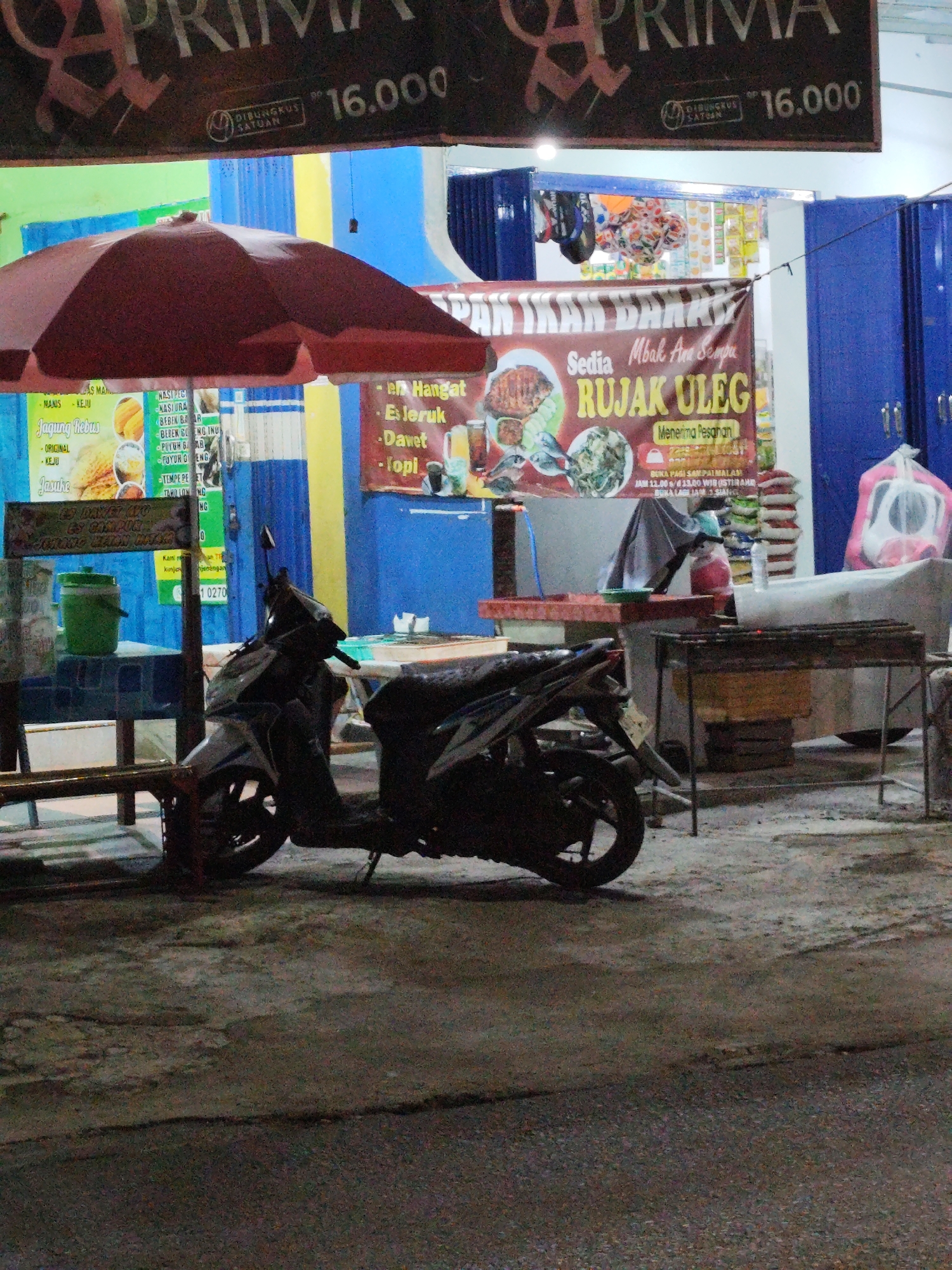 An outdoor scene at dusk or night in the city of Sempu, Indonesia, depicts a food stall with various offerings. A large maroon umbrella shades a food preparation area. A banner advertising "RUJAK ULEG" is prominently displayed. In the foreground, a dark-colored motorcycle is parked. Behind the motorcycle, a charcoal grill is set up, with sacks of what appear to be rice or other dry goods stacked nearby. Blue cabinets form the backdrop of the stall. Signs for various food items, including "JAGUNG REBUS" (boiled corn) and "ES DAWET AYU", are visible. A green bucket sits on a table. The ground is paved with asphalt. The overall impression is that of a typical street food vendor's setup.