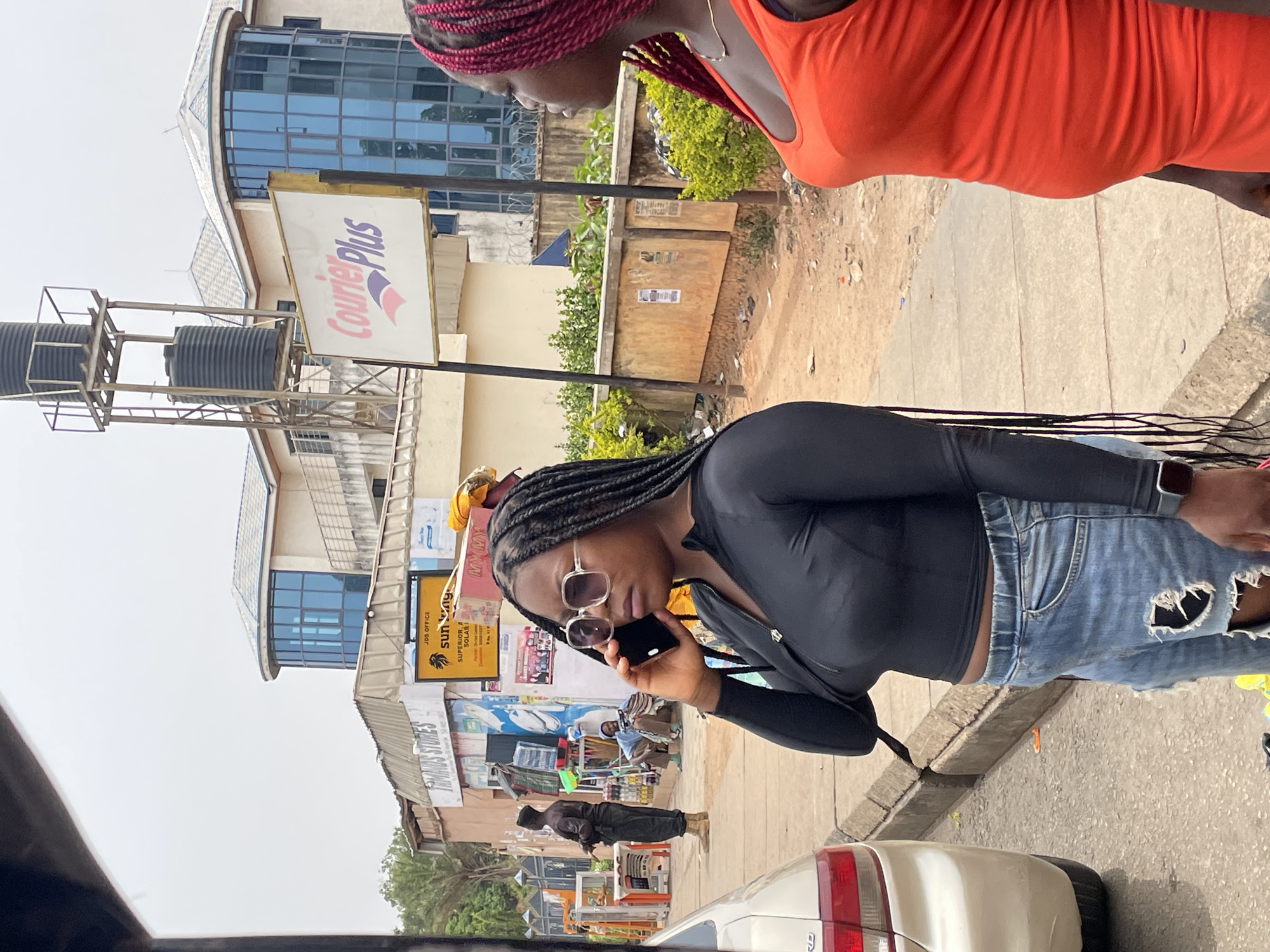 The image captures a vibrant street scene in what appears to be a commercial district in Jos, Nigeria, during the daytime under clear skies. The perspective is slightly angled, looking down a street bustling with activity.
In the immediate foreground, two women are visible. On the right, a woman with red braided hair and an orange dress is partially seen, facing away from the viewer. To her left, another woman, with dark braided hair, sunglasses, a black long-sleeved top, and ripped light-wash jeans, is centrally featured. She is holding a phone to her ear, seemingly engaged in a conversation.
Behind these individuals, the street is lined with various buildings and businesses. A prominent multi-story building features a distinctive curved section with blue-tinted windows. Attached to a structure in front of this building, a sign clearly displays "CourierPlus" in purple and pink text on a white background. Below this, two large black water tanks are mounted on a metal frame.
Further down the street, other commercial establishments are visible. A yellow sign prominently features "SUN RAY" with a lion logo, along with "JDS OFFICE" and partially visible text like "SUPERIOR SOLAR". Another sign below it reads "MIRAGE STORES." Several other individuals can be seen in the background, including a man standing further down the street and another person seated near a shop. Various items are displayed outside these shops, suggesting retail activity.
A white car is partially visible in the lower right corner of the frame. The ground consists of concrete sidewalks and dirt patches, indicating a typical urban setting. The bright, clear sky suggests sunny weather.