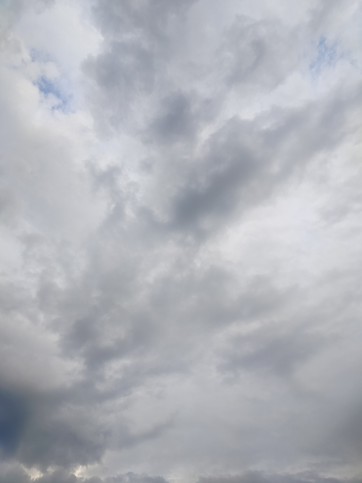 The image is a close-up, low-angle shot of a cloudy sky. The clouds are a mix of light gray and dark gray, with some patches of bright white where sunlight is breaking through. There is a small patch of blue sky visible in the upper left quadrant of the image. The clouds are textured and appear dense, suggesting a potentially overcast or stormy day. There are no discernible features of the city of Дзешчанскі сельскі Савет, Belarus, in this image, as it exclusively depicts the sky.