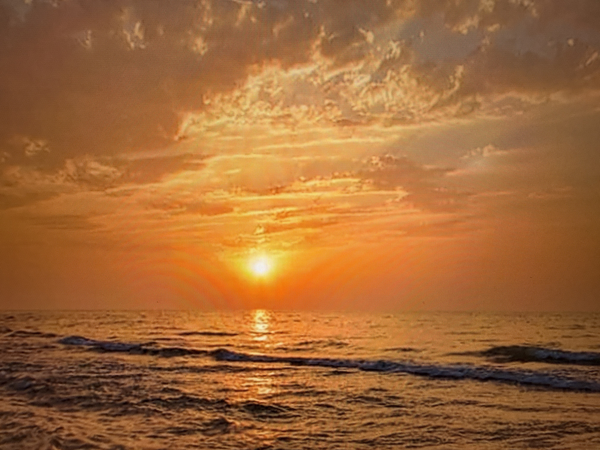 The image depicts a sunset over a body of water. The sky is a vibrant orange, with scattered clouds illuminated by the setting sun. The sun itself is a bright orb low on the horizon, casting a shimmering reflection on the water. Gentle waves are breaking on the surface of the water, creating streaks of white foam. The scene is a natural landscape, and there is no indication of any urban environment or specific location within Szczecin, Poland. No people or specific objects are visible in the image.