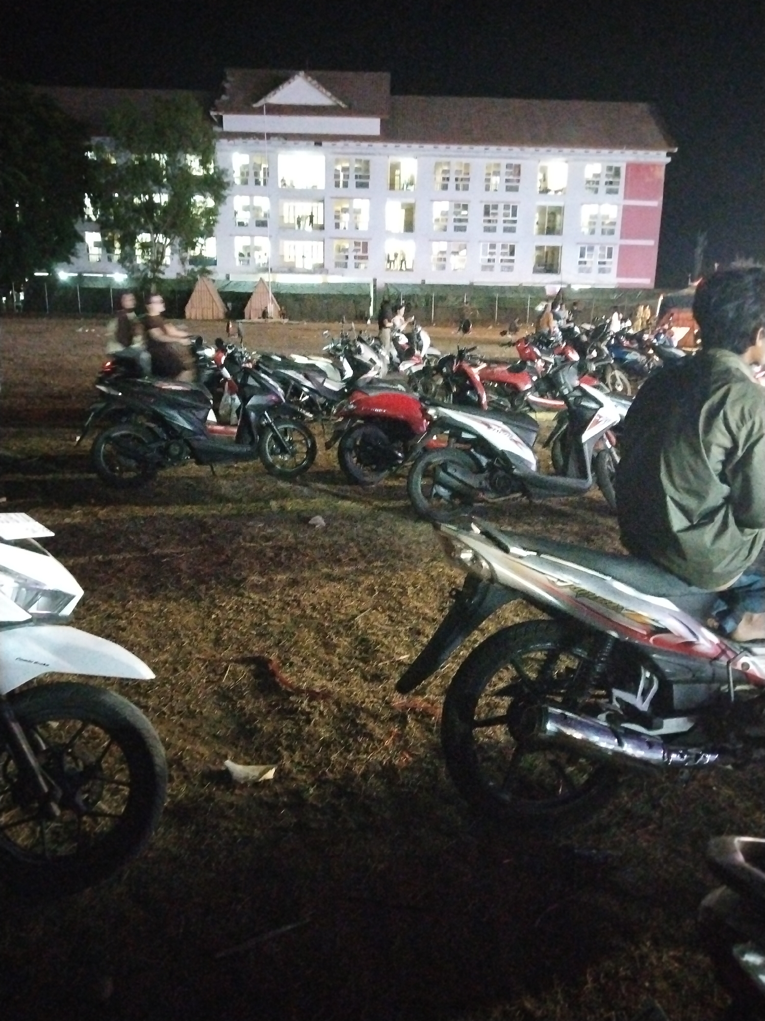 The image depicts an outdoor scene at night, likely in the city of Tasikmadu, Indonesia. In the background, a large, multi-story building with numerous lit windows is visible. In front of the building, a large open area is filled with many parked motorcycles and scooters. Several people are present, some standing and interacting, while others are seated on or near the motorcycles. The ground is covered in dry grass. In the foreground, a white motorcycle is partially visible on the left, and another motorcycle with a rider is in the center right of the frame, with the rider facing away from the camera. The overall impression is that of a gathering or event where motorcycles are a primary mode of transport.