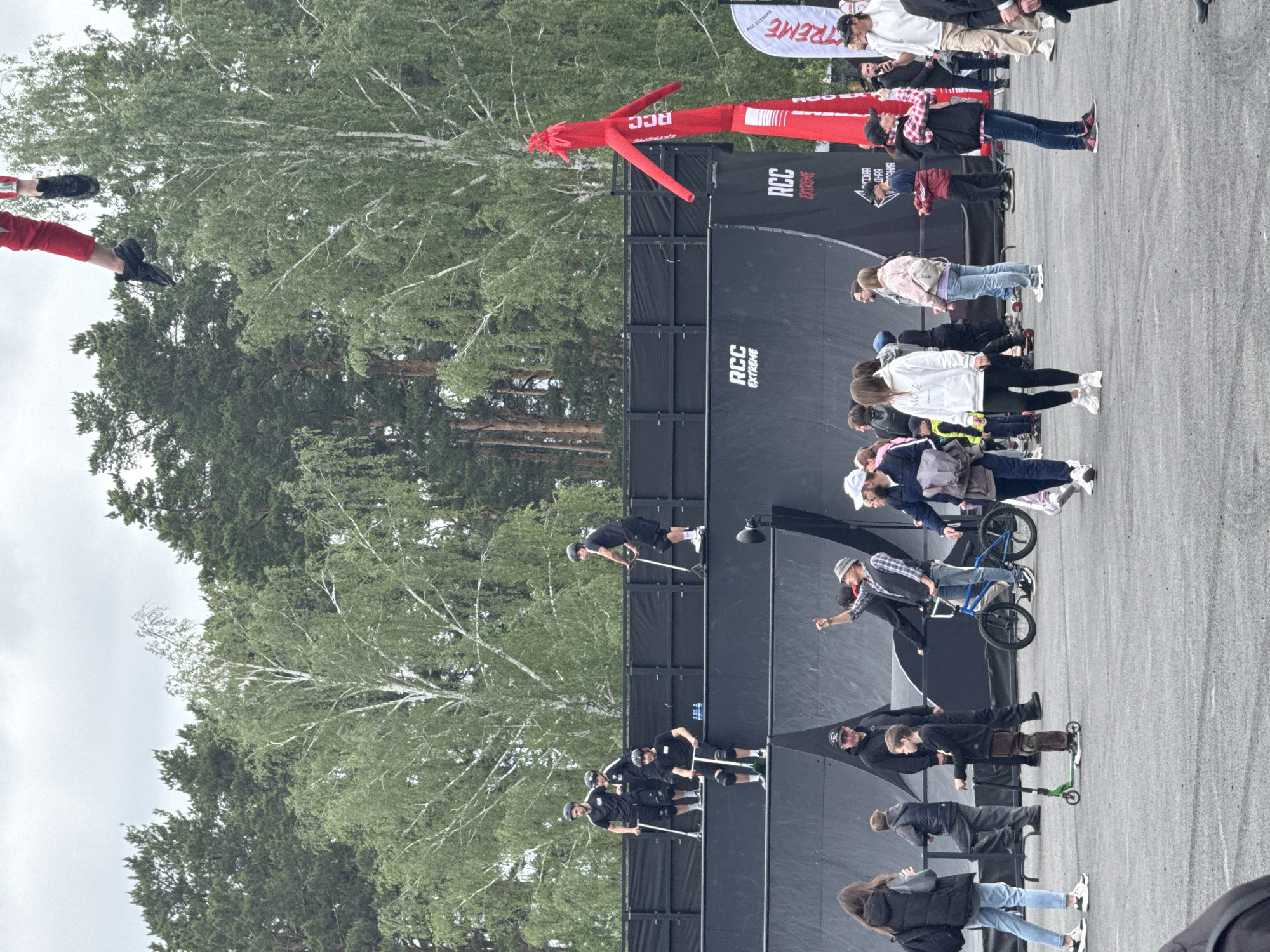 The image shows an outdoor event sponsored by "RCC Extreme," likely in Yekaterinburg, Russia. The event features a large vertical ramp or wall, with a red inflatable tube man positioned above it. A group of people, including several athletes on scooters and a bicycle, are gathered around the ramp. Spectators are watching the activities. Trees and a cloudy sky are visible in the background.