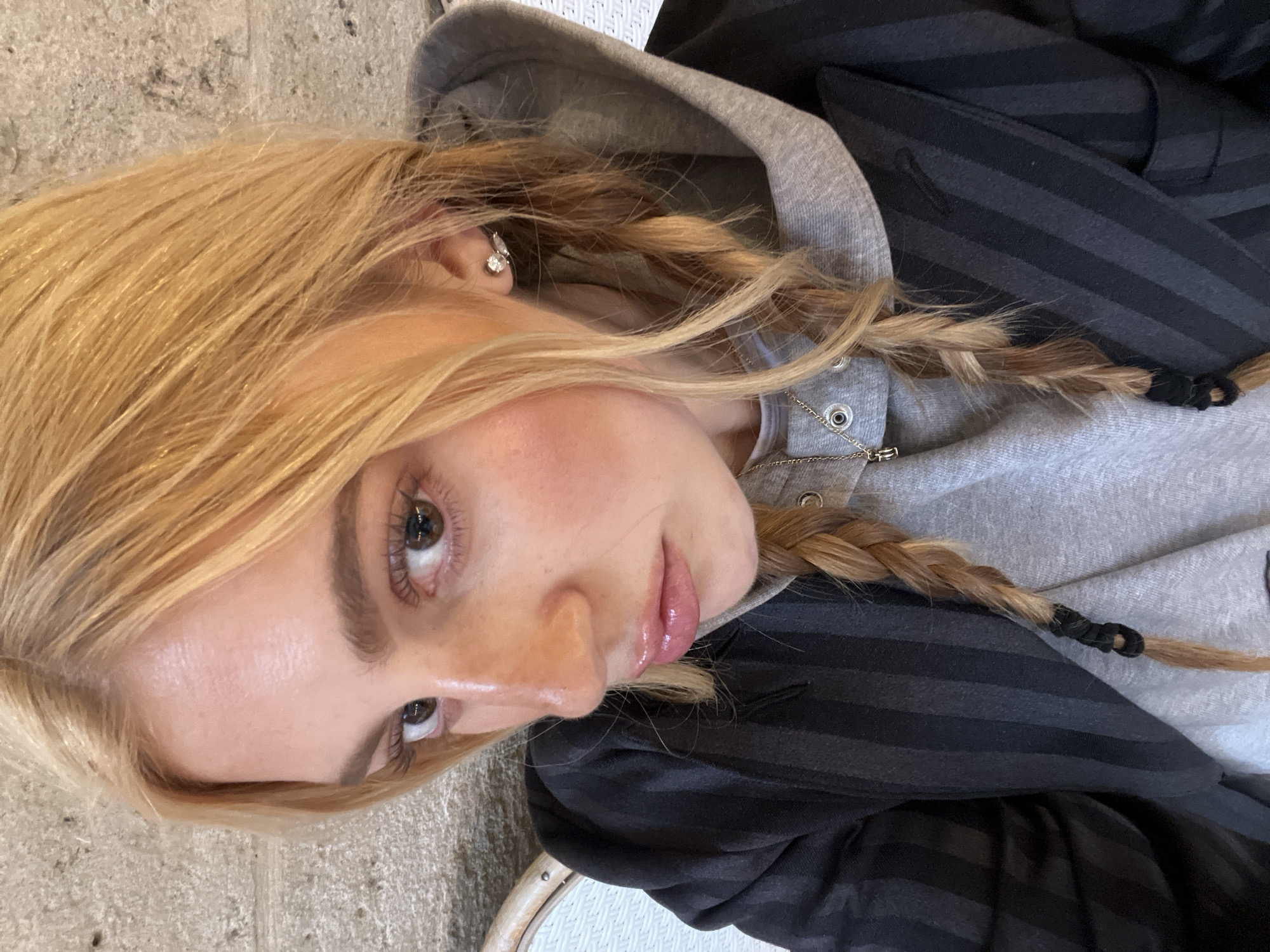 A young woman with blonde hair braided into pigtails looks at the camera. She is wearing a grey hooded sweatshirt and a black and grey striped blazer. She also has a silver earring in her ear. The setting appears to be indoors, possibly a café, but there is no indication that the photo was taken in Paris, France.