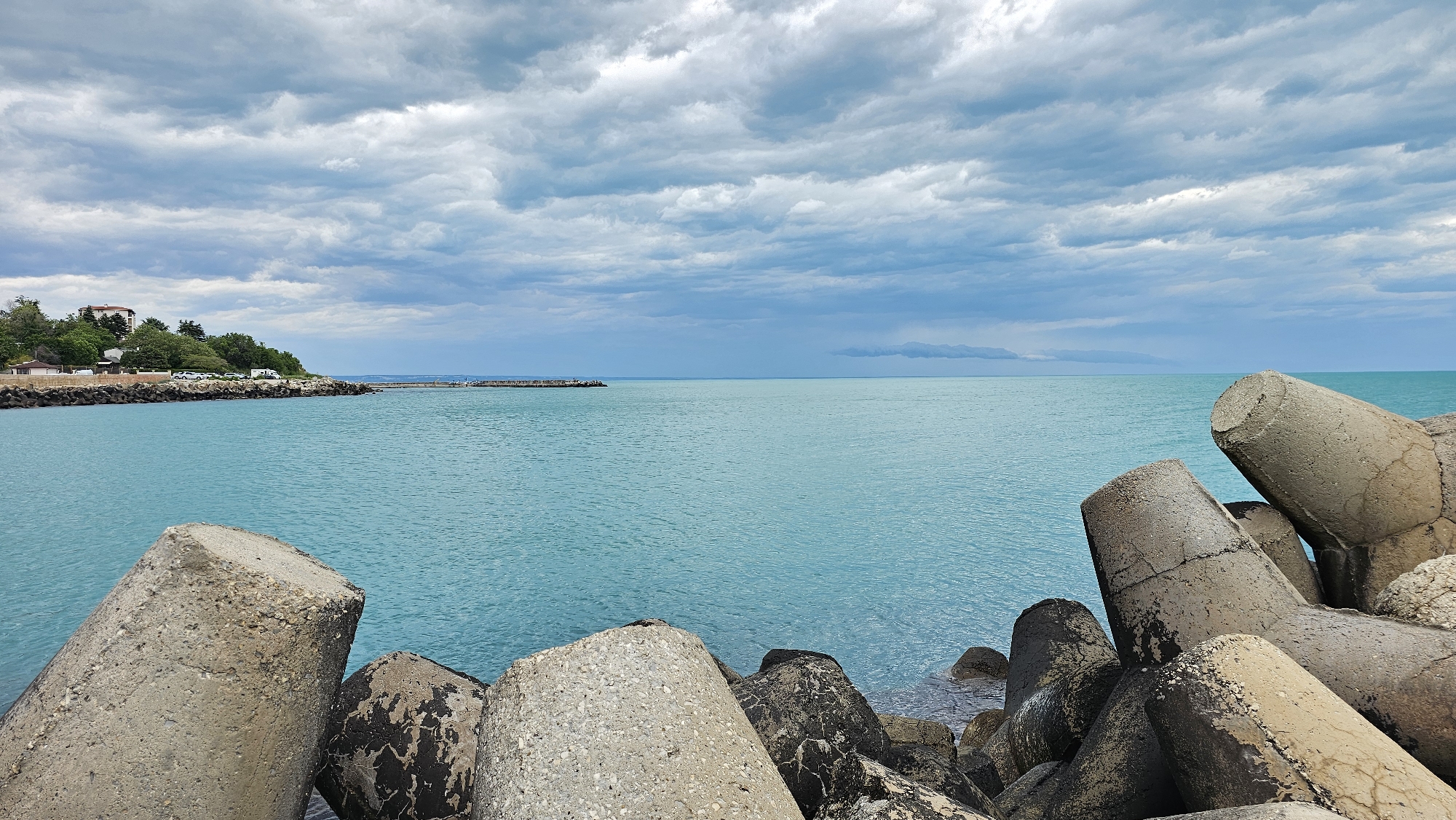 The image depicts a coastal scene in Varna, Bulgaria, characterized by a tranquil aqua sea and a sky filled with textured, cloudy formations. In the foreground, a collection of large, cylindrical concrete tetrapods forms a breakwater, their surfaces weathered and marked with cracks and discoloration. These tetrapods range in color from light gray to darker, almost black, shades due to varying degrees of exposure and weathering.
In the midground, the calm sea stretches towards the horizon under a partly overcast sky. A faint, misty area suggests a possible rain shower in the distance. The coastline to the left features a green area with trees and what appears to be a small structure or buildings, possibly a residential area. A pier or breakwater extends out into the sea from this coastal stretch.
The image captures the typical coastal defense structures and the peaceful yet moody atmospheric conditions common along the Black Sea coast in Varna. There are no people visible in the image. The primary subjects are the tetrapods, the sea, and the sky, with their interactions creating the overall coastal landscape.