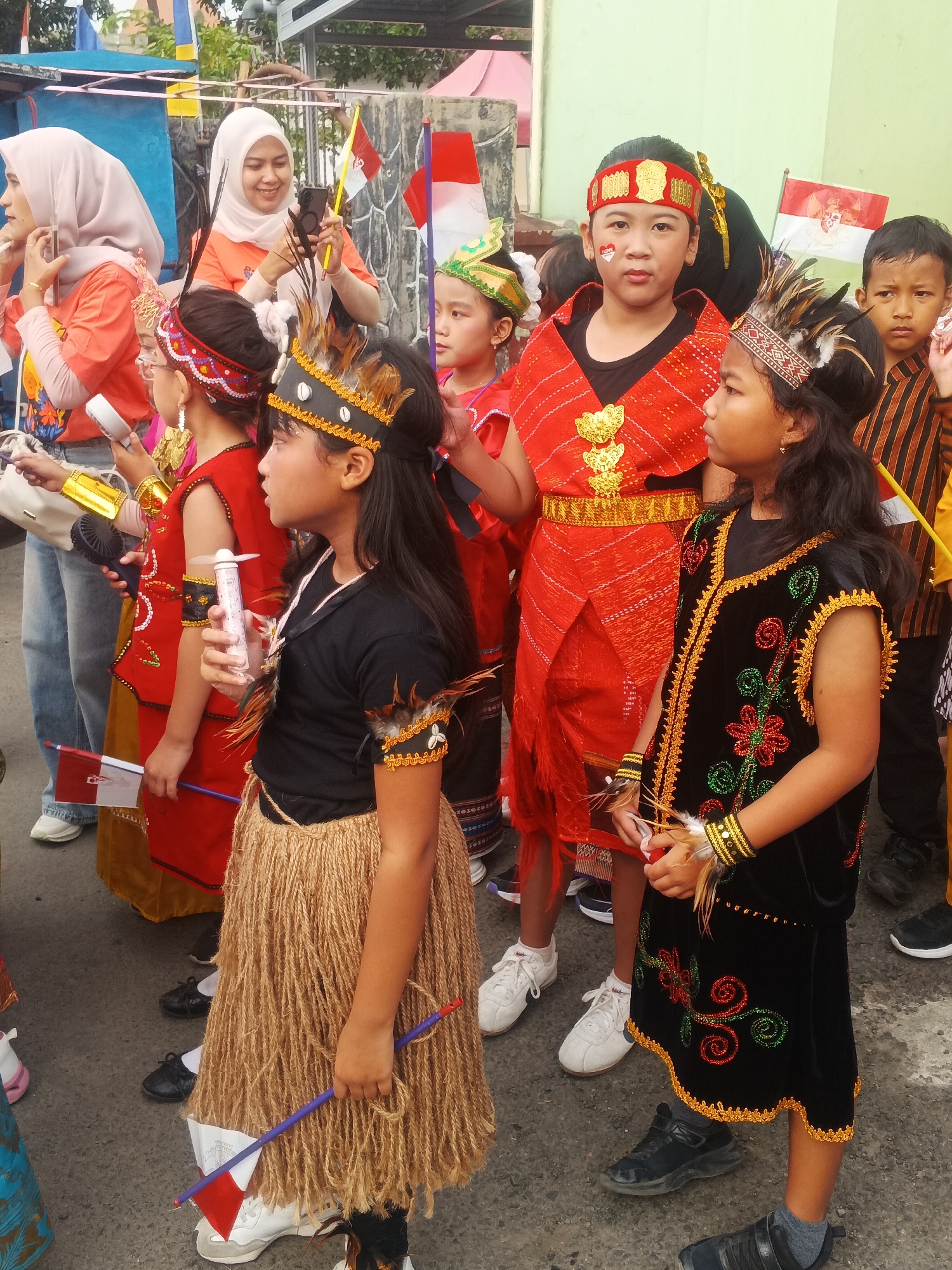 A group of Indonesian children and adults are gathered on a paved street in Sokanegara, Indonesia, possibly for a parade or cultural event. The children are dressed in colorful traditional clothing, some adorned with headbands and feathers. The woman in the foreground, wearing a light pink hijab and an orange shirt, is holding a mobile phone and a small Indonesian flag. Another child in the foreground is holding a similar flag. The background shows buildings and trees, typical of an urban Indonesian setting.