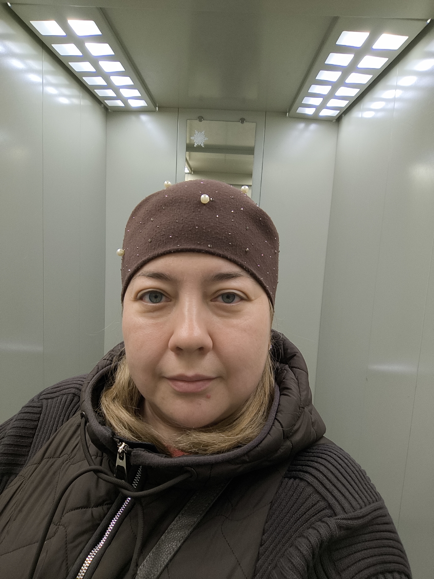 A medium-shot selfie, taken at eye level, depicts a Caucasian woman with blonde hair and blue eyes wearing a brown beanie adorned with pearls and a dark brown quilted vest with a ribbed sweater underneath. The woman's expression is neutral. The background reveals an elevator interior with white walls, overhead fluorescent lights, and a small mirror containing a snowflake decoration. The location is Khimki, Russia.