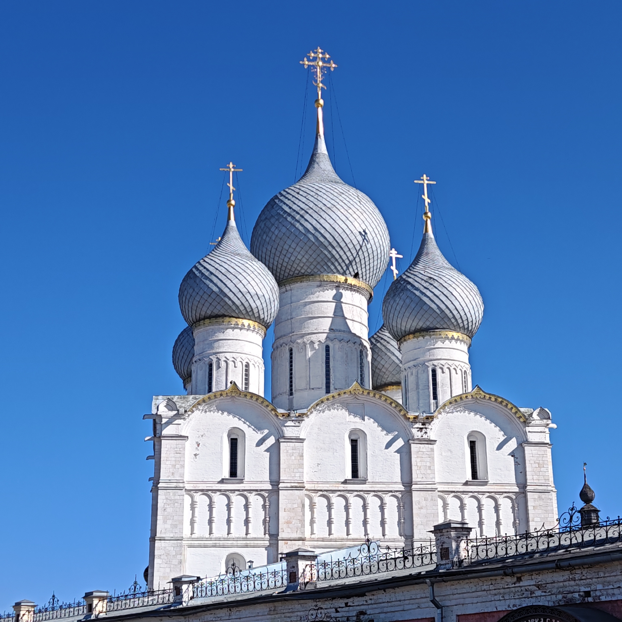 The image shows the Dormition Cathedral in Rostov, Russia, with its distinctive onion domes and gold crosses against a clear blue sky. The cathedral's white facade is visible, displaying its architectural details such as windows and decorative arches. A wrought-iron fence and part of a building are visible at the bottom of the image in front of the church, but there are no people visible in the image.