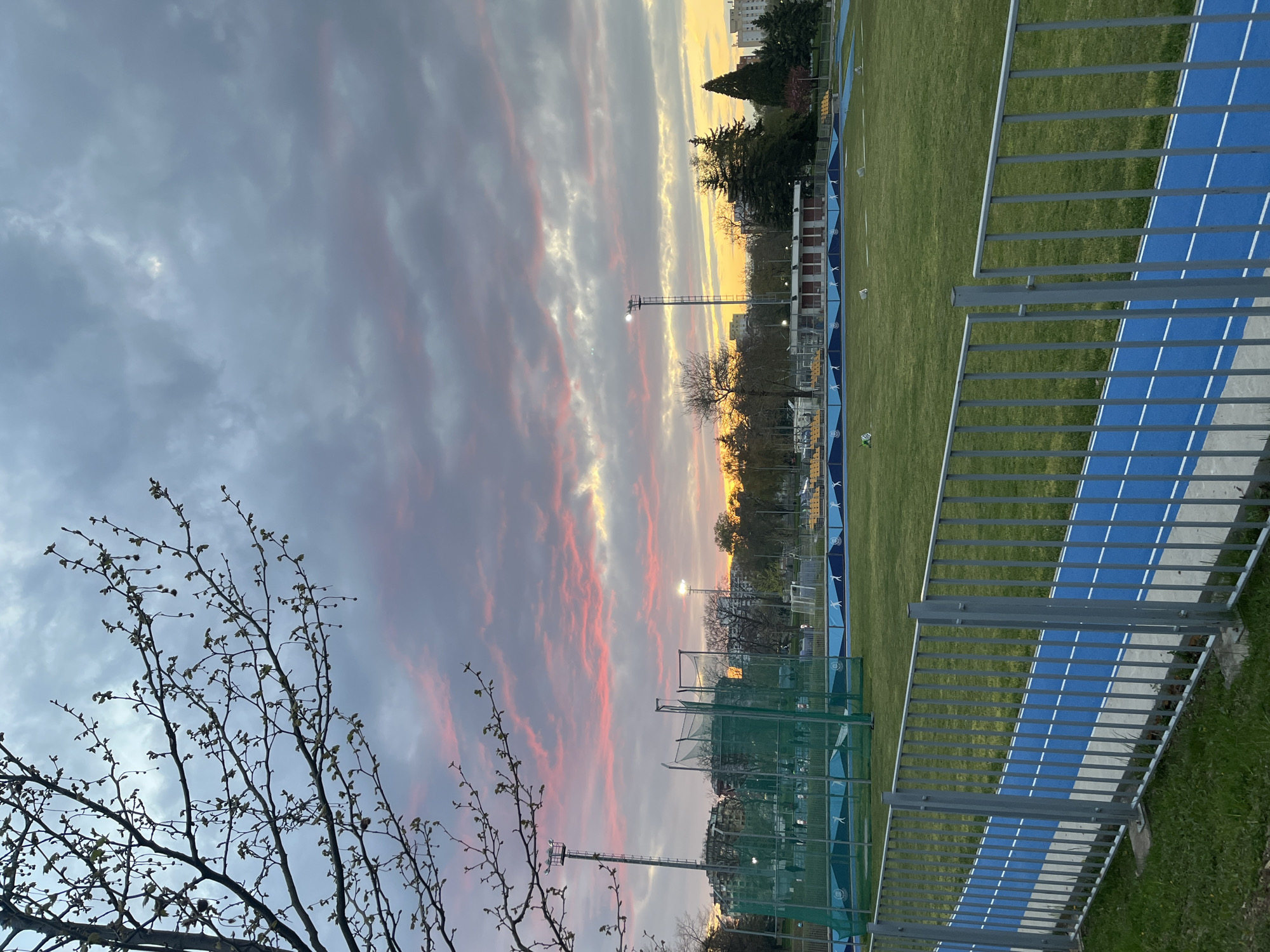This media file captures an outdoor sports complex during either sunrise or sunset, evidenced by the dramatic sky. The image is oriented sideways but depicts a horizon line with the sky at the top and the sports grounds below.
The sky is a prominent feature, dominated by a mix of dark grey clouds, with vibrant streaks of orange, pink, and yellow light near the horizon, indicating the low position of the sun. In the foreground on the left, the bare branches of a tree with budding leaves are silhouetted against the colorful sky.
The scene features a green grassy sports field, likely a soccer or football pitch, bordered by a blue running track with distinct white lane markings. A tall, gray metal fence separates the viewer from the track and field. Another green net fence is visible closer to the field. Several tall floodlight poles, one of which is illuminated, stand around the perimeter of the field, suggesting evening or early morning use. A small white soccer ball is visible on the grass.
In the mid-ground, beyond the track, there are evergreen trees and a few bare deciduous trees, with urban or suburban buildings visible in the distance. A blue barrier or low fence runs along the edge of the field, decorated with repeated white logos or stylized figures, possibly depicting athletes, though no specific text is legible.
No people or active events are visible, lending a quiet and serene atmosphere to the scene. The time of day adds a beautiful, tranquil quality to the sports venue.