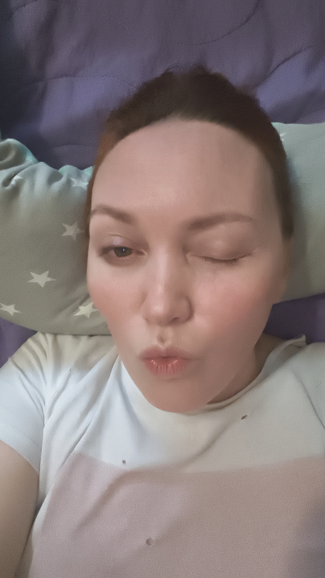 A woman with auburn hair is lying down, partially supported by a pillow with white star patterns. She is wearing a white t-shirt with a light pink horizontal stripe at the bottom. One eye is closed in a wink, and her lips are pursed in a kiss. The background consists of a purple sheet or blanket. The location is not specified and is marked as Unknown, Unknown.