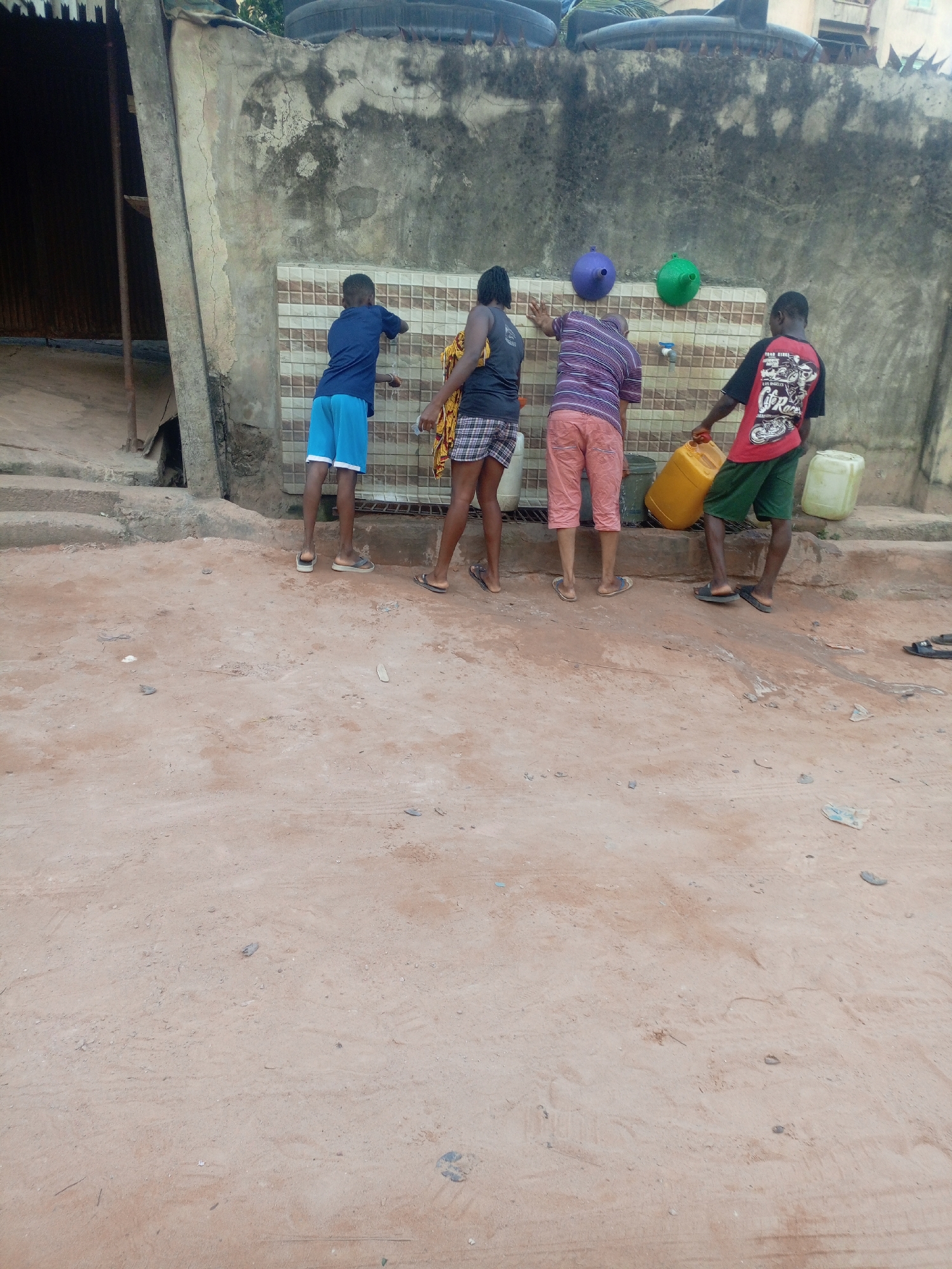 The image depicts an outdoor scene in Onitsha, Nigeria, where four individuals are gathered at a public water collection point, likely during the daytime. The ground in the foreground is unpaved, consisting of reddish-brown earth with some scattered debris and tire marks.
The primary subjects are four people, appearing to be children or young adults, standing in a line facing a weathered concrete wall. A section of this wall, at about hip height, is covered with light-colored, square tiles and features a single blue water tap. Above the tiled section, two brightly colored, funnel-shaped objects – one purple and one green – are affixed to the wall.
From left to right:
1. A young boy wearing a blue t-shirt and blue shorts stands closest to the left, facing the wall.
2. The second person wears a dark tank top and checkered shorts.
3. The third individual wears a horizontally striped t-shirt (purple, white, and black) and light-colored shorts, positioned near the tap.
4. The fourth person, on the far right, wears a red t-shirt with black sleeves and green shorts. This individual is actively holding a large yellow jerrican, seemingly in the process of filling it with water. The red t-shirt has visible text which reads "ROAD RIDERS" and below it, "G.P. Race" or "Off Road" with graphics. A white jerrican is also placed on the ground near this person.
In the background, atop the concrete wall, several large black water tanks are visible, suggesting the water source for the taps. To the far left, a partial view of a corrugated metal structure and a large wooden beam indicates adjacent buildings or a sheltered area. The scene portrays a common daily activity of communal water collection.
