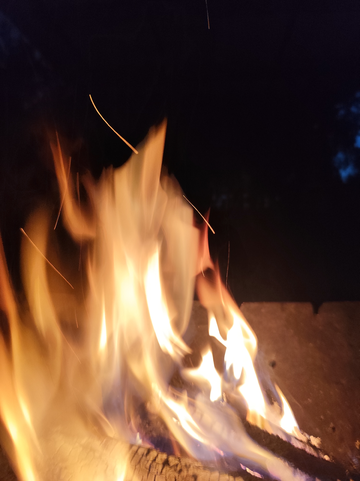 The image depicts a close-up view of a campfire at night. The primary subject is the vibrant, dynamic flames of the fire, which fill most of the frame with oranges, yellows, and white hues. Several glowing embers and sparks are visible, appearing as thin, bright lines against the dark background, indicating movement within the flames. The fire is burning on what appears to be logs, with one log clearly visible in the foreground, showing texture and charring. In the background, a dark, blurry area suggests the night sky and possibly some distant foliage or trees, with a hint of blue light that could be moonlight or ambient light. The overall setting is outdoors at night, likely in a natural or semi-natural environment, given the presence of a campfire. There is no indication of people, and the focus is entirely on the fire itself. The mood evoked is one of warmth and natural energy, typical of a campfire scene. There is no visible text in the image. The time of day is clearly night.