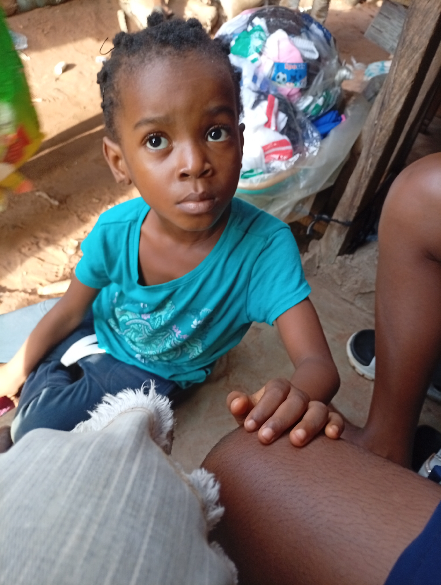 A young girl with dark skin and braided hair looks directly at the camera with a neutral expression. She is wearing a turquoise t-shirt with a leafy pattern and dark pants. She is sitting on the ground, and her left hand rests on her knee, while her right hand is placed on the thigh of another person whose dark legs are visible in the foreground. Beside the girl, on the right side of the frame, a bundle of clothes wrapped in plastic is visible, with wooden planks behind it, suggesting an outdoor setting in an informal environment. The ground appears to be dirt, and the lighting suggests it is daytime. The overall scene is likely in Nkpor, Nigeria.