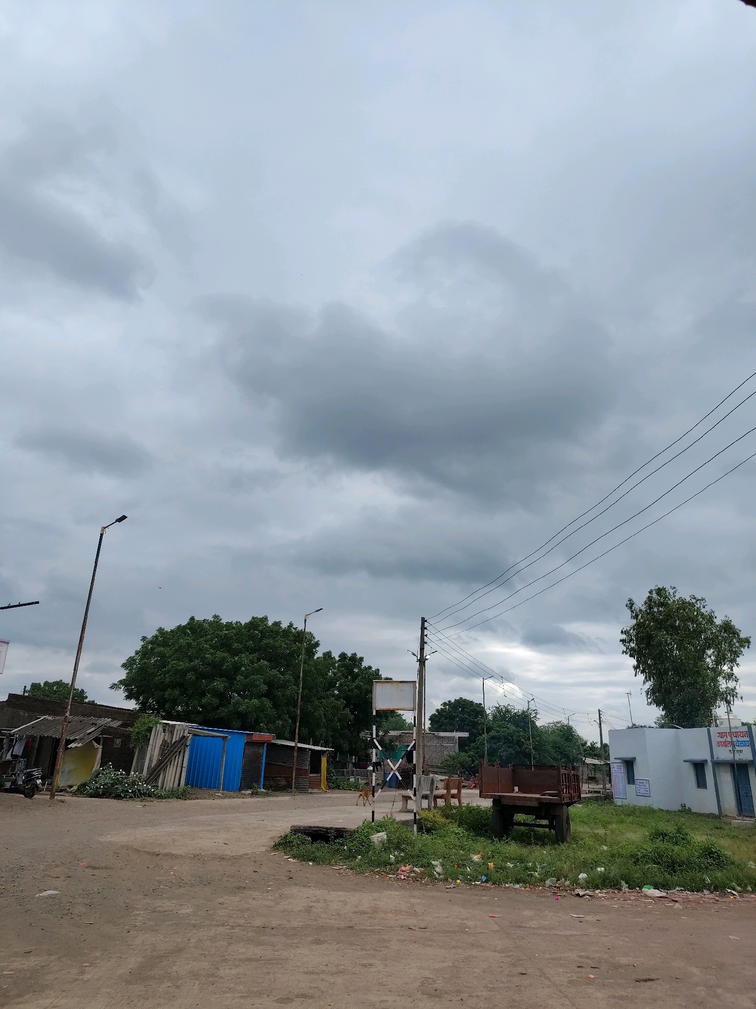 This image captures a street scene in Velawad, India, under an overcast sky with dark, heavy clouds. The foreground features a dirt road and a grassy area littered with debris. To the left, a cluster of ramshackle structures with corrugated metal roofs and blue siding are visible, adjacent to a parked scooter. A tall, single-arm street lamp stands near these structures. Further into the midground, a rusty trailer is parked on the grass. Behind it, a small, white building with blue trim bears signage that appears to be in Hindi. Several trees, including a large one with dense foliage, are scattered throughout the scene. Power lines stretch across the upper portion of the frame, supported by utility poles. A second, taller street lamp is visible further down the road to the right. The overall impression is one of a rural or semi-urban environment on a cloudy day.