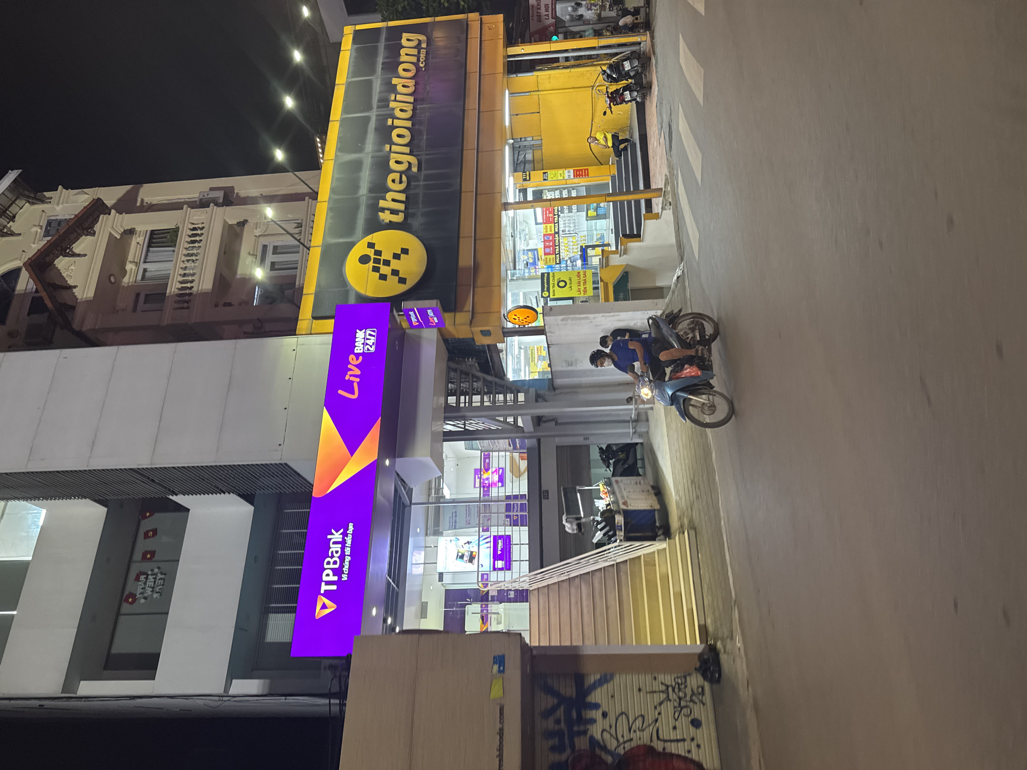 The image depicts a street scene at night in Hanoi, Vietnam, specifically in front of two adjacent businesses: TPBank and The Gioi Di Dong. The TPBank branch, with its prominent purple signage, is located to the left, featuring the bank's logo and Vietnamese text. To its right is The Gioi Di Dong, a mobile phone retailer, identified by its bright yellow facade and signage with the store's name and a pixelated logo.
A person is seen riding a blue motorbike in the foreground, moving from left to right across the street. The motorbike has a slight blur, indicating movement. The street itself is paved and appears wet, reflecting the ambient light from the businesses. Pedestrian crossings are visible on the road.
The architecture of the buildings is multi-story, with the TPBank and The Gioi Di Dong occupying the ground floors. A traditional-looking building with balconies and ornate detailing stands to the right of The Gioi Di Dong, illuminated by streetlights. A few parked motorbikes are visible near the entrance of The Gioi Di Dong. The scene is dimly lit by streetlights and the neon glow of the business signs, creating a nocturnal urban atmosphere. Graffiti is present on a rolled-down metal shutter to the far left of the frame.