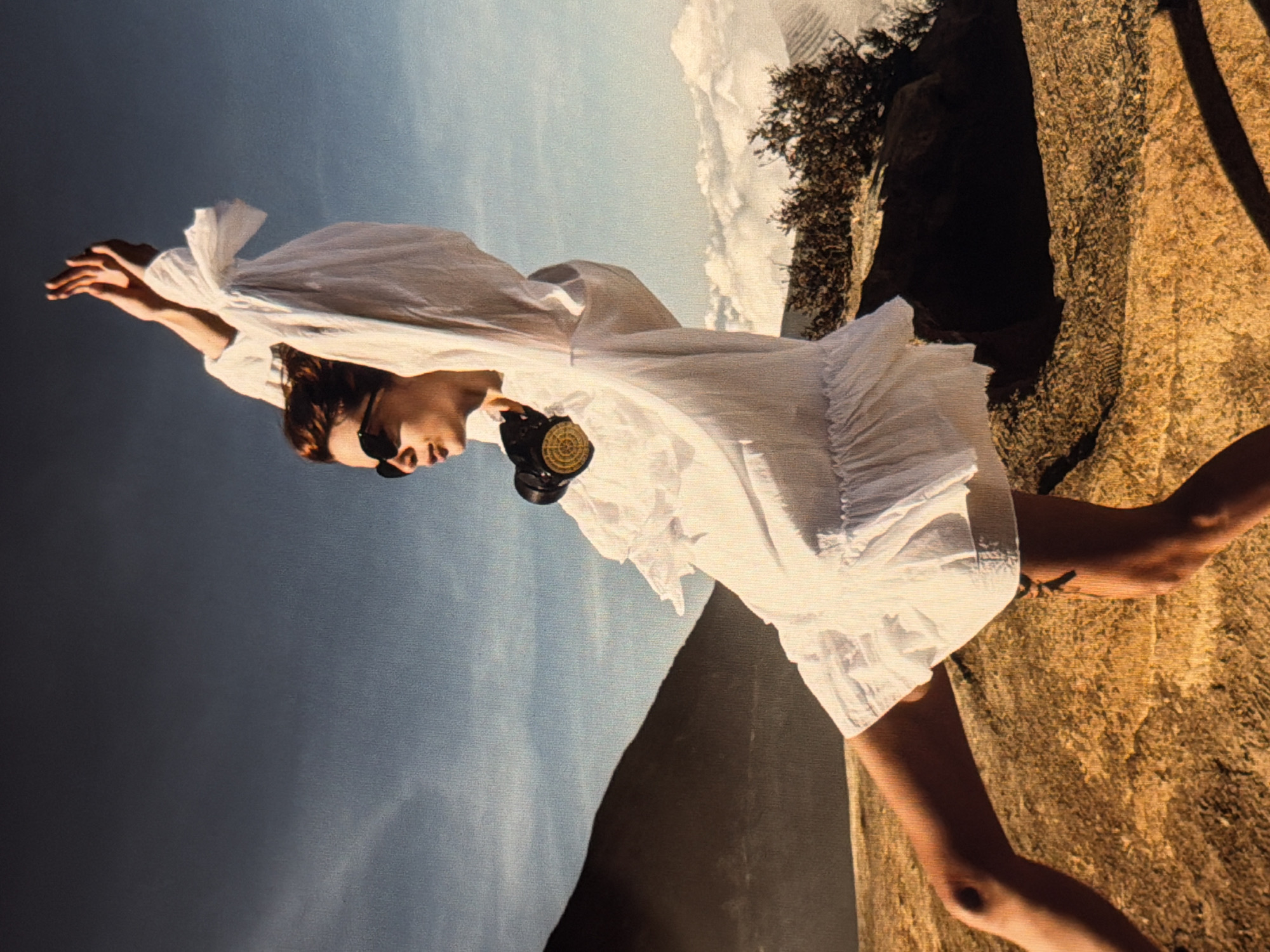 A woman in a short white dress stands on a rocky mountain outcrop. She wears sunglasses and a large, dark-colored object (possibly a camera) hangs around her neck. Her arms are raised, with the fabric of her dress flowing around her. To her left, a mountain slope drops away into a misty, bluish sky. To her right, the rocky outcrop is interspersed with some vegetation. The background includes sky and clouds with rock and vegetation on a mountain. I cannot determine if the image is of Đà Nẵng, Vietnam.