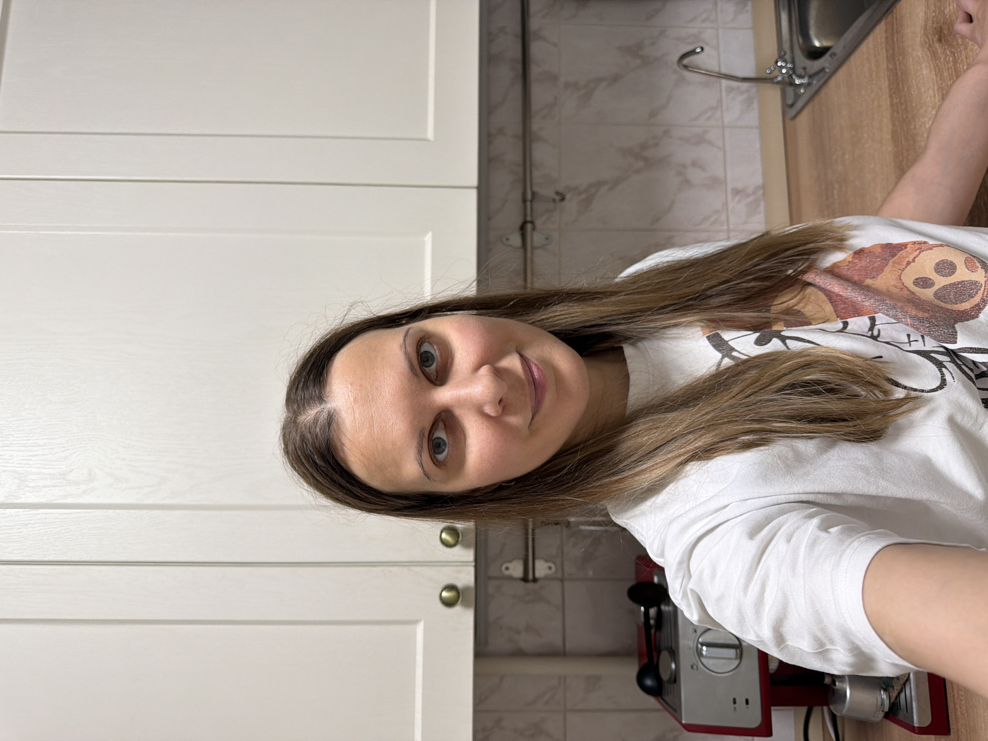 A person is holding a smartphone at head-level, in selfie orientation, in front of a kitchen setting. The subject appears to be a female with brown hair, wearing a white t-shirt with a graphic print. Her face is visible, showing light skin and light-colored eyes.
Behind her are light-colored kitchen cabinets with gold-colored knobs. The countertop is wood-toned, and there is a stainless steel sink and faucet visible. The backsplash has a light, marbled tile pattern. An espresso machine is visible behind her arm.
The location is likely inside a residential kitchen; the location in the city of Izhevsk, Russia, is unknown based on the provided image. No specific actions or interactions are apparent, and only one person is visible.