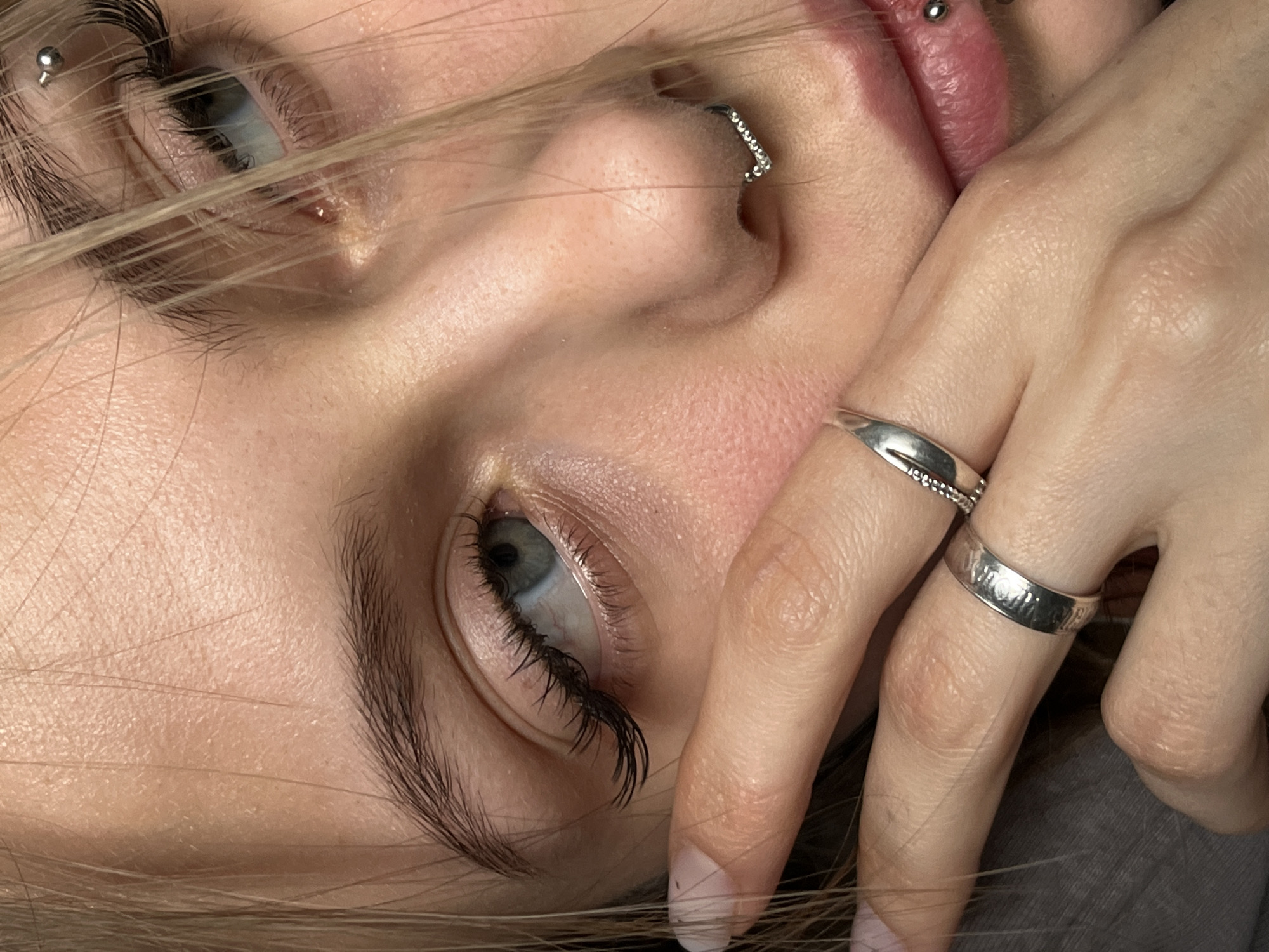A close-up shot presents a young woman's face at an oblique angle. Her skin has a fair complexion, and the fine blonde hair is partially obscuring her face. Both eyes are visible, revealing light-colored irises surrounded by dark lashes. The woman sports multiple piercings: one above her left eyebrow, a bejeweled ring in her left nostril, and a small stud on her upper lip. Her right hand is raised, with two silver rings adorning her fingers.
The image does not provide any information that indicates a location in Фоки, Russia.