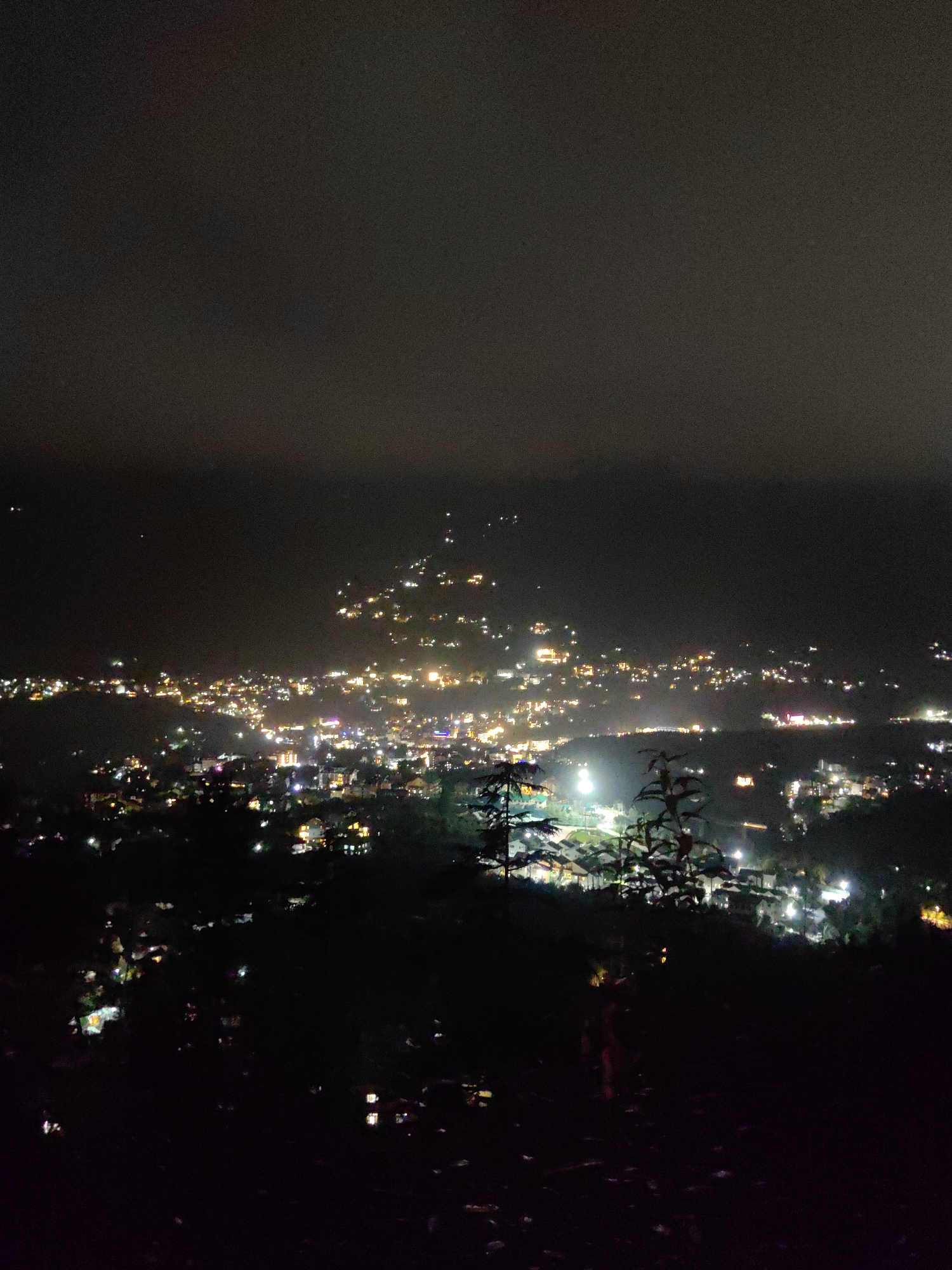 A high-angle, night-time shot captures a sprawling cityscape illuminated by numerous lights. The urban area is nestled within a valley, with buildings densely packed along rolling hills. The sky above is dark and overcast, with a few scattered lights visible in the distance, possibly stars or distant aircraft. The foreground is dominated by the dark silhouettes of trees and foliage, creating a sense of depth and framing the illuminated city below. The city itself displays a variety of light colors, including white, yellow, and pink, suggesting different types of lighting and structures. The overall impression is of a vibrant, active city seen from a distance at night. The location is identified as Siumsa, India.