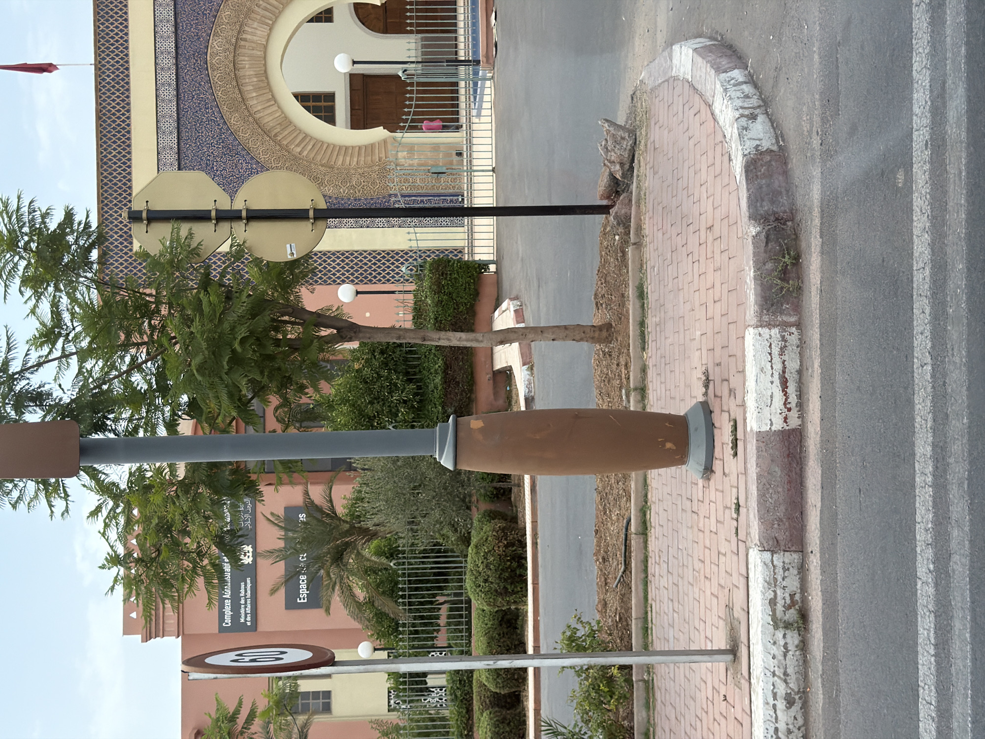 The image captures a street scene in Marrakesh, Morocco, focusing on the exterior of a building with distinctive Moroccan architectural elements. A prominent feature is a tall, brown, cylindrical lamppost with a dark gray pole, situated on a paved walkway. To the left of the lamppost, a white pole supports two signs: a circular sign with "60" in white on a red background, indicating a speed limit, and a yellow octagonal sign above it.
Behind the lamppost and signs, the building presents a facade decorated with intricate geometric patterns in blue, gold, and white. A large, arched entryway is visible, with wooden doors. The building's exterior is painted in shades of salmon and terracotta. Green metal fencing encloses an area in front of the building, with manicured bushes and trees providing greenery. A red Moroccan flag flies from a flagpole on the roof of the building.
The ground in the foreground consists of a paved street with white road markings. The walkway is made of red brick pavers bordered by concrete curbs. The lighting suggests it is daytime, with overcast skies. The scene appears to be an entrance to a public building or complex.