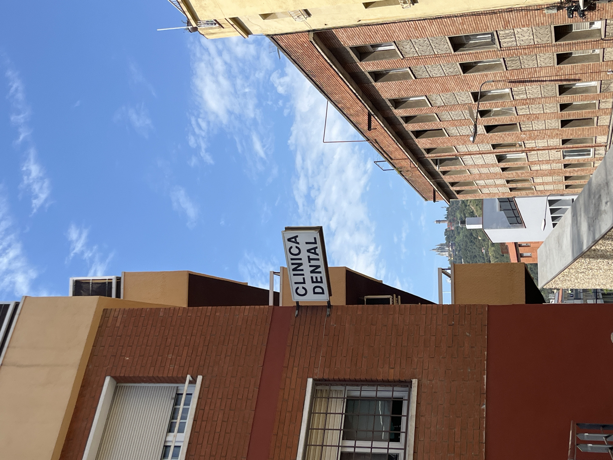The image presents an upward-looking perspective of urban buildings under a bright blue sky with scattered white clouds, suggesting clear weather on a sunny day. The overall scene is static, devoid of people or specific events.
In the immediate foreground, a building with a reddish-brown brick facade is prominent. This building features sections of smooth, brownish-cream colored plaster alongside the brickwork. Two windows are visible: one with a white rolling shutter partially lowered, and another secured by a dark metal grille. Perched atop this building is a white rectangular sign with the clear inscription "CLINICA DENTAL" (Dental Clinic) in black capital letters.
To the right and appearing taller, another building with a similar reddish-brown brick facade extends upwards. Its design incorporates a grid-like pattern of numerous windows framed by the brickwork and alternating grey, possibly concrete, panels. Further in the background, a section of a light yellow building is visible. Through a gap between the structures, distant green hills or mountains, dotted with subtle indications of more buildings, can be seen, providing a geographical context that aligns with Barcelona, Spain. Various wires and utility fixtures are also discernible against the sky and attached to the buildings.