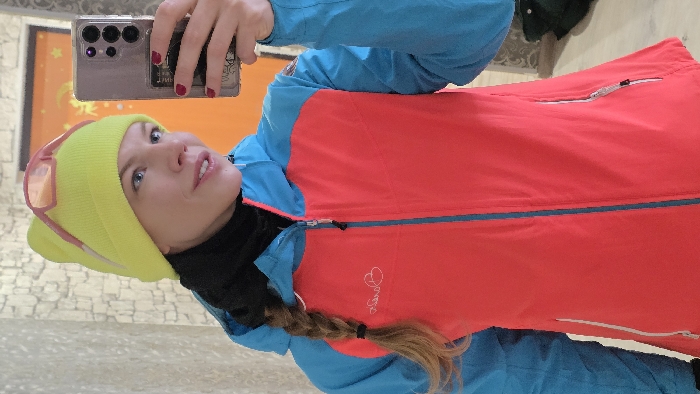 A woman is captured in a selfie, holding a smartphone with her right hand to take the picture. She is dressed in cold-weather attire, suggesting an outdoor or cool indoor setting. Her face is turned slightly upwards, looking towards the phone's camera, with a neutral to slightly curious expression. She has light-colored eyes, possibly blue or grey.
She is wearing a bright neon yellow beanie that covers her ears, and a pair of pink-framed sunglasses are perched on top of the beanie. Beneath the beanie, a black neck gaiter or balaclava is visible around her neck and lower face. She is wearing a vibrant red vest or jacket over a blue long-sleeved jacket or shirt, the sleeves of which are visible. Her hair, which appears to be light brown or reddish-brown, is styled in a braid that extends over her right shoulder.
The smartphone she is holding appears to be a modern device with multiple camera lenses, possibly a Samsung Galaxy S23 Ultra, encased in a clear protective cover. Her fingernails are painted a dark red or maroon color.
The background of the image is somewhat out of focus. Behind her head and upper body, there's a large, brightly lit area that appears to be an orange-colored wall or a section of a painting with some lighter yellow star-like shapes. Below that, the wall features a textured pattern resembling light-colored stacked stones or bricks. The overall impression is that the photo was taken indoors, possibly in a changing room, a ski lodge, or a similar casual setting before or after an outdoor activity. There is a small, light-colored embroidered logo on the red vest, which seems to include a stylized heart shape and some illegible text next to it.