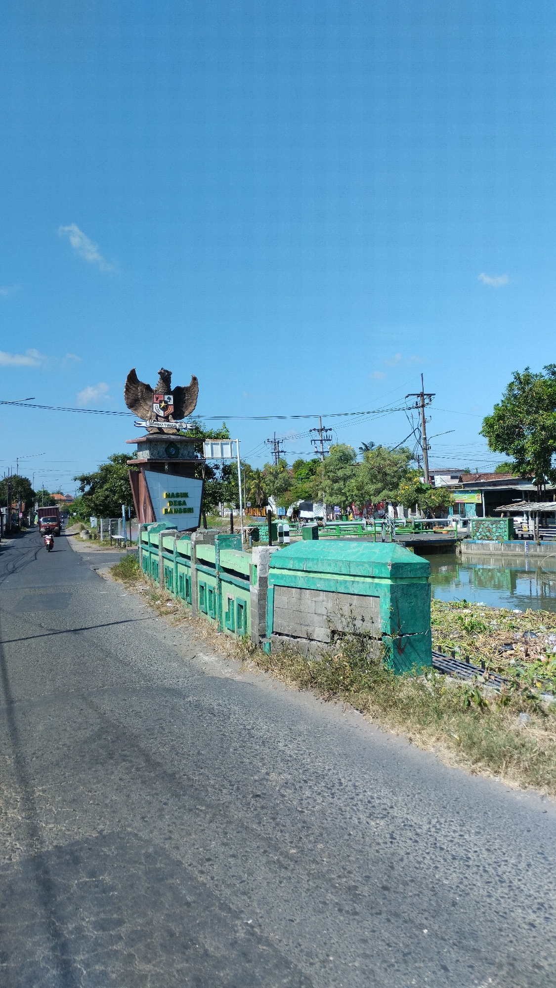 The image captures a street scene in Plumbungan, Indonesia, dominated by a large monument featuring the Garuda Pancasila, the national emblem of Indonesia. The monument is positioned on the left side of the road, with text below it that appears to read "PASUK DESA BAMB..." which likely signifies a village name. To the right of the monument, a canal or river runs parallel to the road, bordered by a green, ornate fence. Lush green trees are visible in the background, interspersed with utility poles and wires. On the road, a motorcycle is in motion, indicating movement. The sky is a clear, bright blue with a few wispy clouds, suggesting a sunny day. The road surface is asphalt, showing some wear and tear. The overall environment appears to be a semi-urban or rural area within Plumbungan.