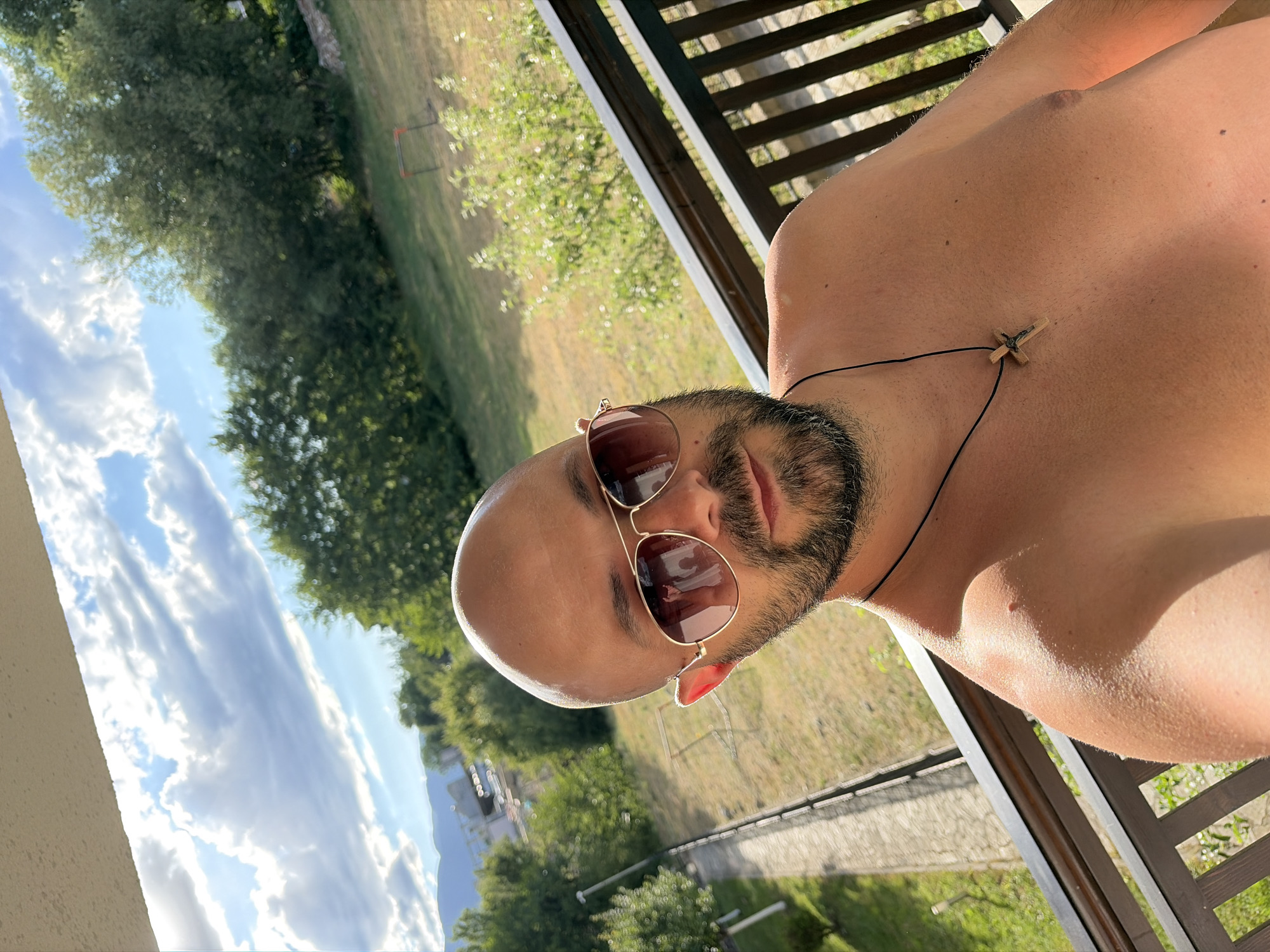 The image is a selfie taken by a shirtless man with a bald head, beard, and sunglasses, standing on a balcony. He is wearing a wooden cross necklace. The background shows trees, a lawn, a path, and mountains in the distance under a cloudy sky. The specific location is a residential area in Bansko, Bulgaria, identifiable by the characteristic Balkan architecture and mountainous landscape typical of the region.