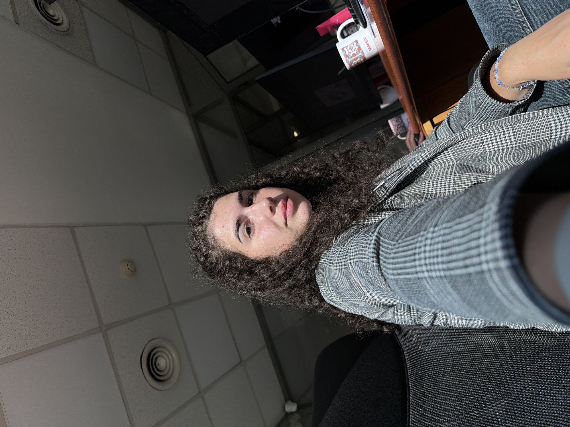 The image shows a woman with curly dark hair sitting in an office chair. She is wearing a grey plaid blazer and blue jeans. Her left arm is resting on the armrest of the chair, and she is wearing a beaded bracelet on her wrist. In the background, there is a wooden desk with a computer monitor, a mug with a floral pattern, and a pink object. The ceiling has acoustic tiles and recessed lighting. The overall lighting suggests the sun is shining through a window, casting shadows. The scene appears to be an office environment in Sofia, Bulgaria.