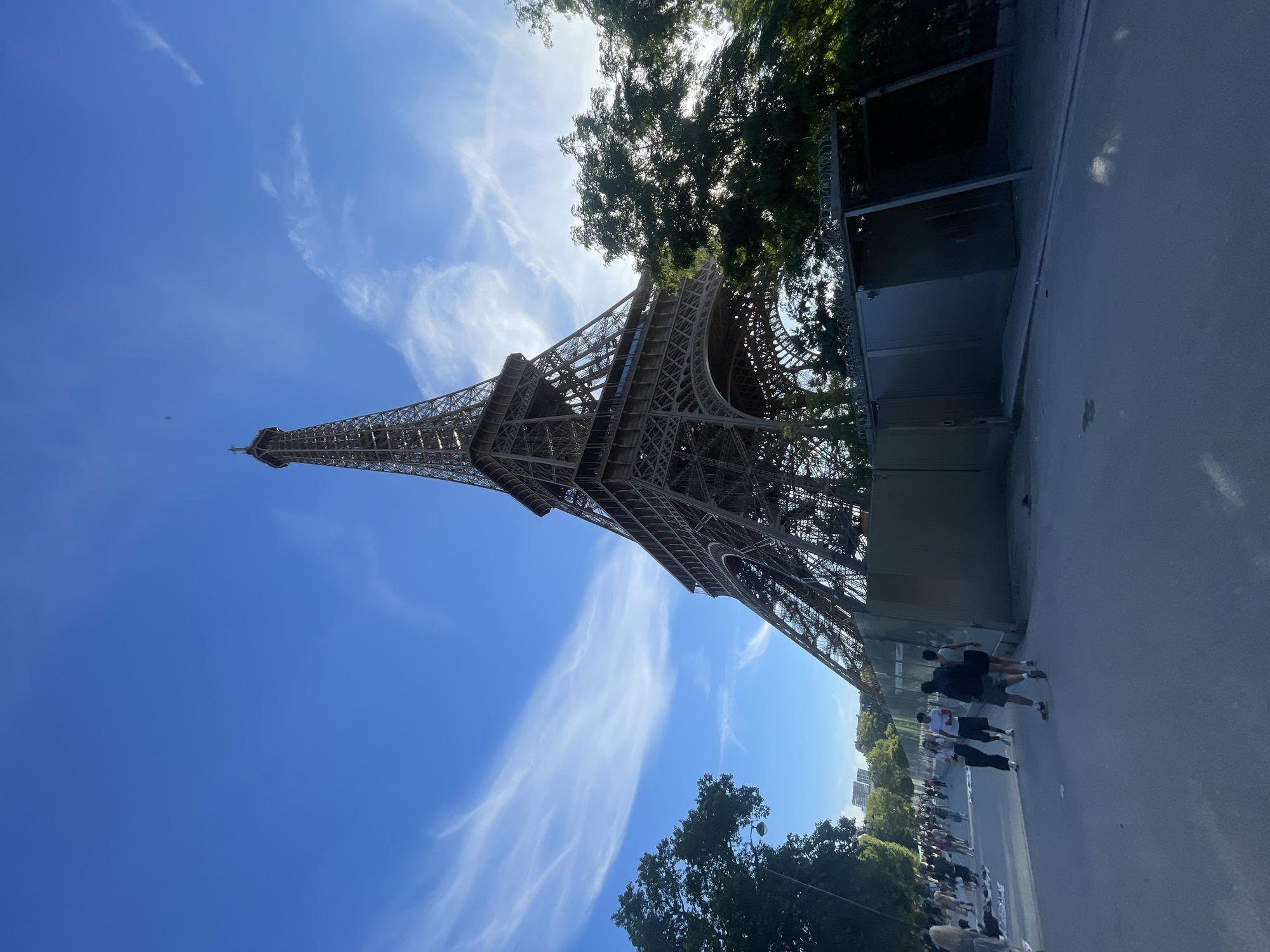The image shows the Eiffel Tower in Paris, France, viewed from a low angle with the sky taking up a significant portion of the frame. Wispy white clouds are scattered across the bright blue sky. The iconic iron lattice structure of the Eiffel Tower dominates the right side of the image, with its peak extending towards the top. In the foreground, a paved pathway is visible, with several people walking along it. Some of these individuals are in small groups, engaged in conversation or walking at a leisurely pace. To the left of the pathway, lush green trees provide a natural element, and a lamppost is visible amongst them. To the right of the pathway, a temporary construction barrier or enclosure is in place, partially obscuring the base of the Eiffel Tower and adorned with barbed wire at the top. The environment appears to be an outdoor public space, likely a park or plaza surrounding the monument.