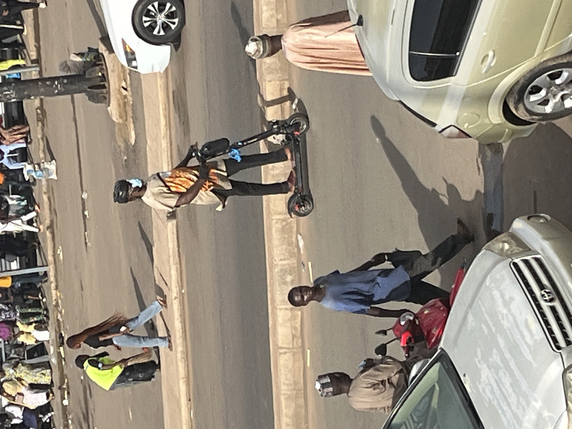 This is an eye-level, medium shot of a street scene in Jos, Nigeria. In the foreground, a silver car is parked on the left, with a red motorcycle to its right. A person wearing a blue shirt and dark pants is walking away from the motorcycle towards the right, carrying a black bag. To the left of the motorcycle, another person in a tan outfit and a patterned cap is seated. Further back, a person is riding an electric scooter, wearing a tan shirt and black pants, with a face mask on. A white car is parked on the right side of the street. In the background, a group of people are standing and sitting. The environment is an urban street with paved roads and a sidewalk.