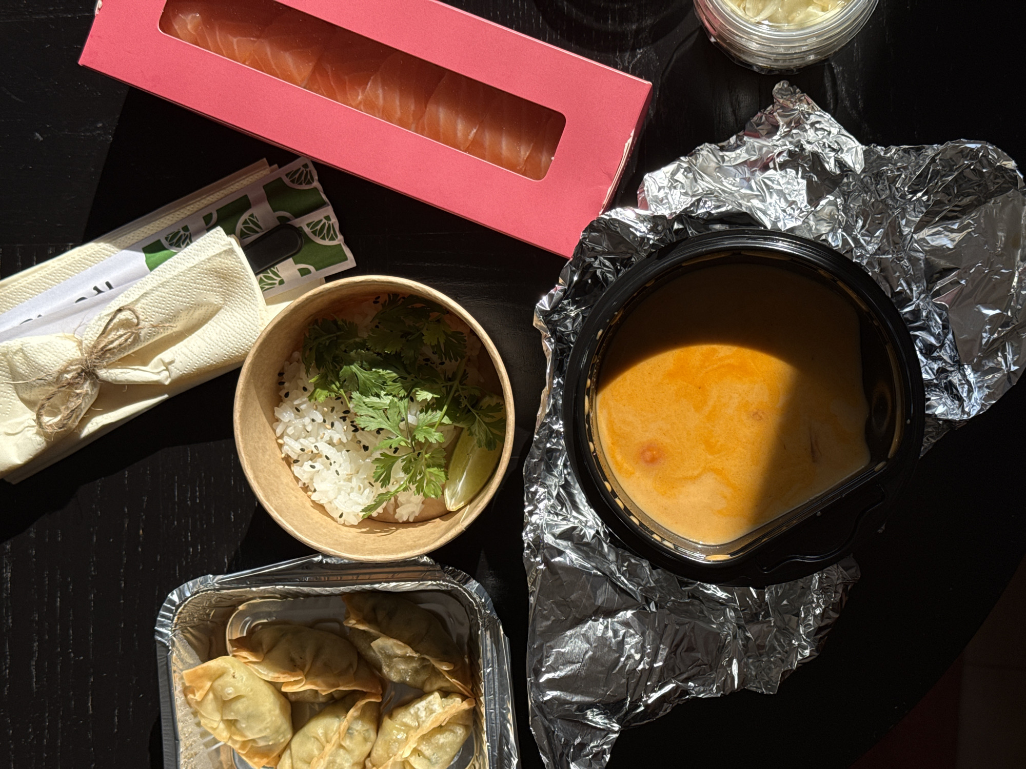 The image shows a table top view of a meal consisting of various Asian-inspired dishes. A salmon nigiri set is in a rectangular pink container, chopsticks in paper packaging, a bowl of rice with herbs and a lime wedge, a container of fried dumplings, and a bowl of Tom Yum soup sitting on aluminum foil. A small container of sauce is also visible. The table is dark, possibly black wood. The location and other information about the city of Vozdovac Urban Municipality, Serbia cannot be determined from the visual context.