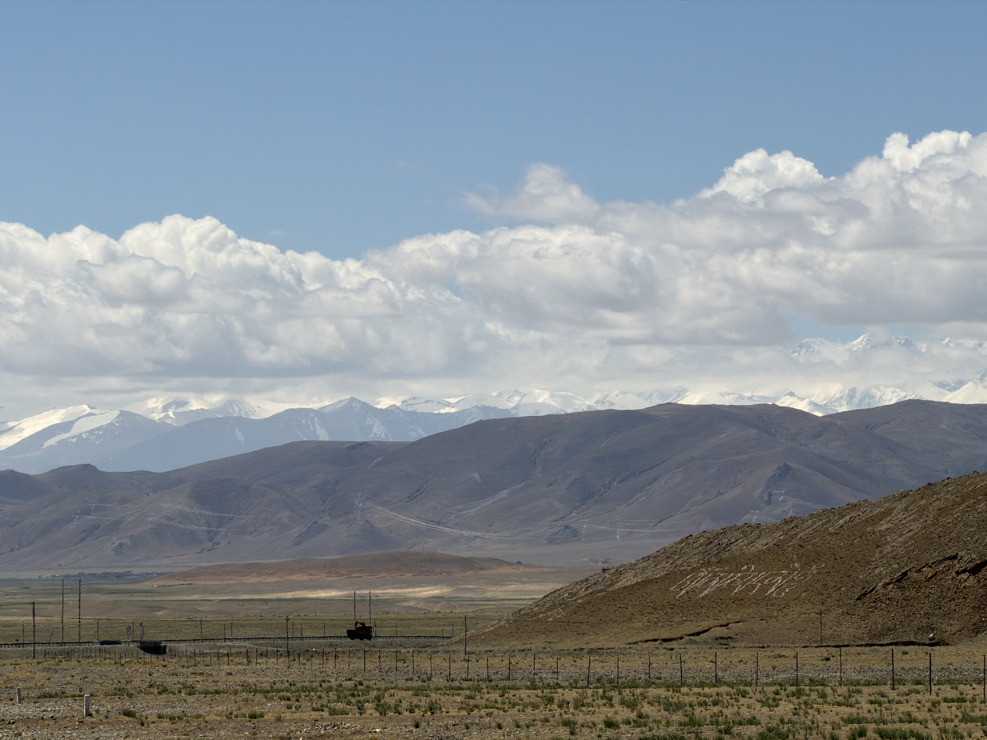 The image presents a vast, expansive landscape in Mainpu township, China, under a bright, partly cloudy sky. The scene depicts a succession of mountain ranges and an arid plain, suggesting a high-altitude or remote region.
In the foreground, the ground is covered with dry, rocky soil and sparse, light-colored vegetation. A multi-strand wire fence runs horizontally across this area.
The midground features a long, straight road or railway line, also bordered by fencing. A small, dark-red or orange truck is visible on this path, moving from left to right. Beyond the road, a wide, flat valley leads to a series of brown, undulating hills. On the right side, a prominent brown hill displays large, light-colored characters formed on its slope, which read "欢迎光临" (Huānyíng guānglín), meaning "Welcome."
In the background, multiple layers of mountains stretch across the horizon, with their peaks becoming progressively fainter. The furthest ranges are distinctly snow-capped, their white summits contrasting with the darker rock.
The sky occupies the upper portion of the image, showing a clear blue with large, fluffy white cumulus clouds forming a distinct band across the middle. The time of day appears to be daytime, with clear visibility and calm weather. No people are visible in the scene, which conveys a sense of grandeur and remoteness.