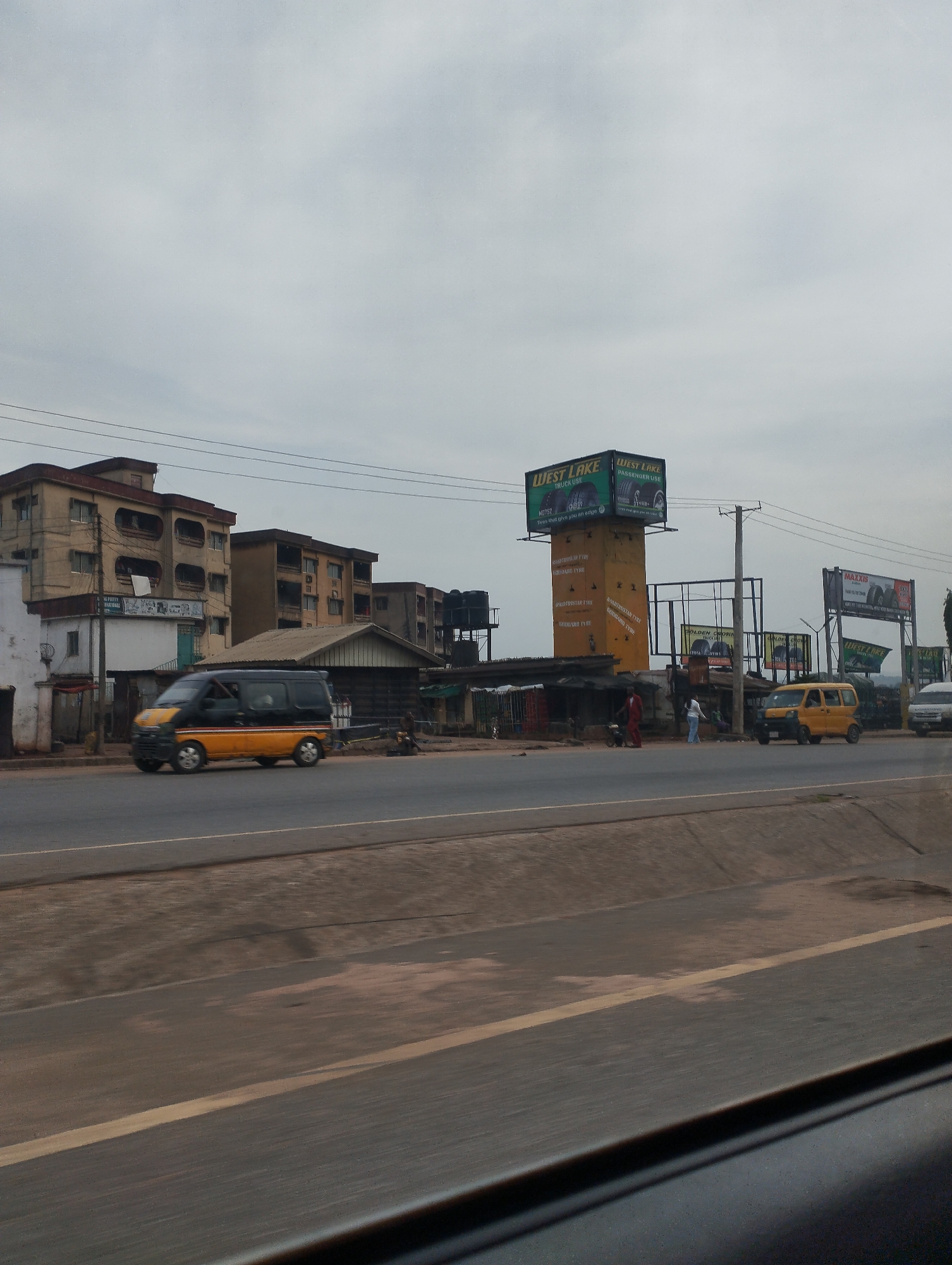 The image presents an urban scene in Ogbunike, Nigeria. A road runs horizontally through the foreground, with a gray and cloudy sky overhead. On the opposite side of the road are several buildings, including multi-story residential structures and smaller shops. Traffic consists of commercial vehicles, including vans painted in yellow and black. A billboard displaying advertisements for 'West Lake' tires and 'Maxxis' tires is visible, indicating a commercial district. There are also some individuals present near the shops, but the image does not reveal any specific actions or interactions.