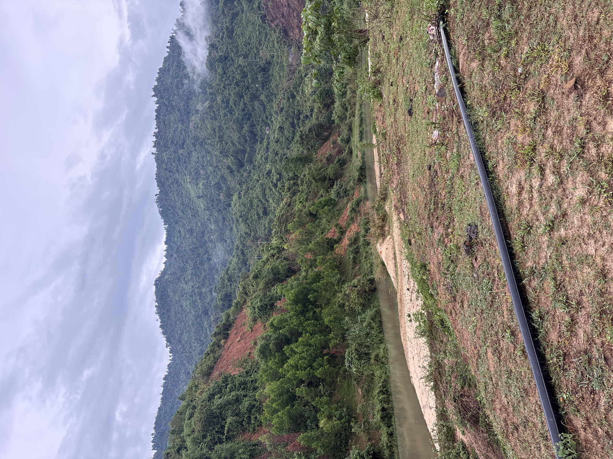 The image shows a landscape in Khanh Vinh, Vietnam, featuring a mountain range partially obscured by clouds. The mountains are densely vegetated with trees, showing signs of deforestation on one slope with exposed reddish soil. A narrow body of water, possibly a river or stream, flows through the landscape. A black pipe lies on the grassy ground. The sky is overcast with gray and white clouds.